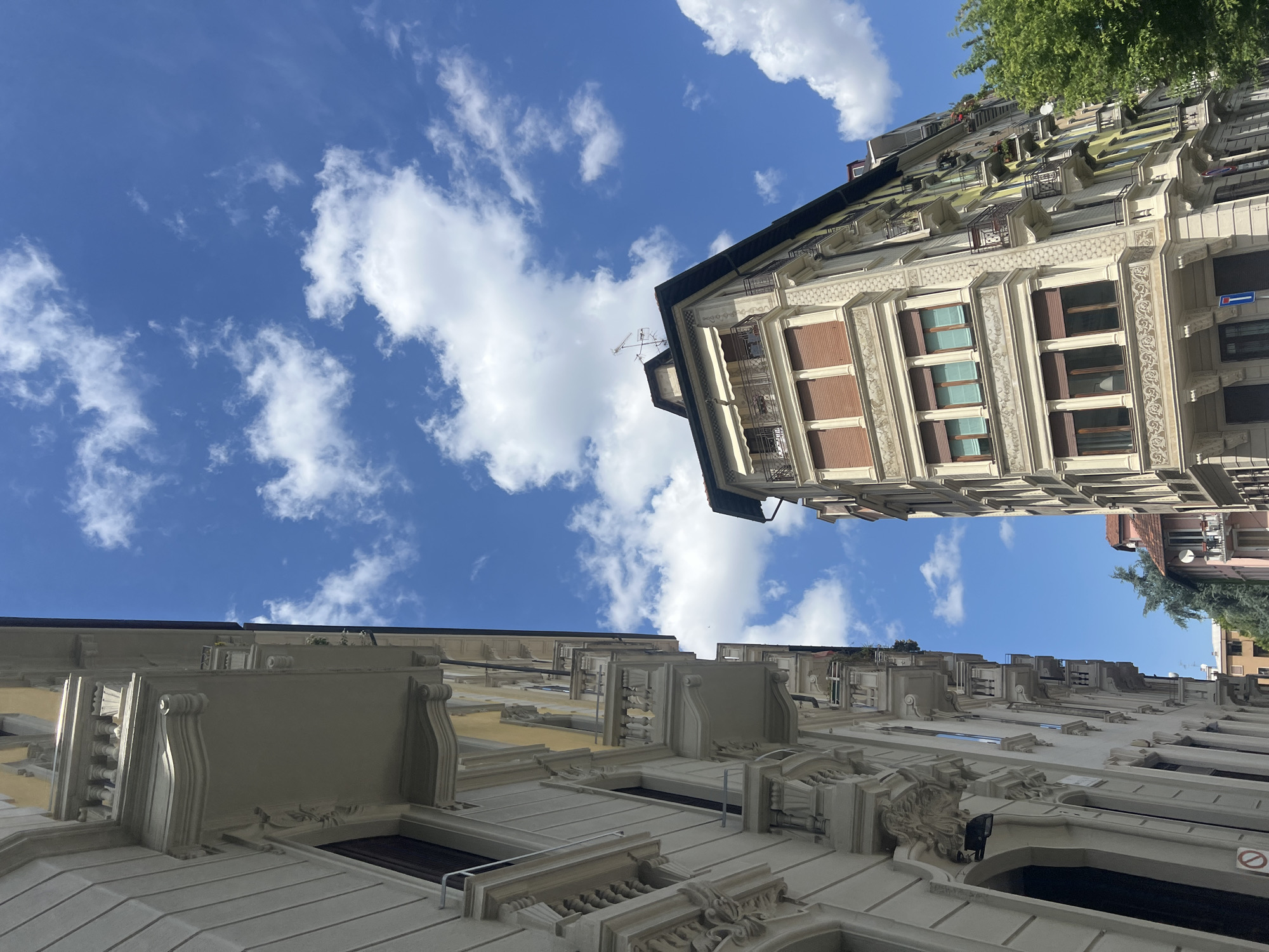 The image presents an upward-angled view of buildings against a bright blue sky dotted with puffy white clouds. The architectural styles of the buildings vary, with one building showing an ornate facade, including elaborate window trim, balconies, and detailed stonework. The buildings are located in the city of Milan, Italy.