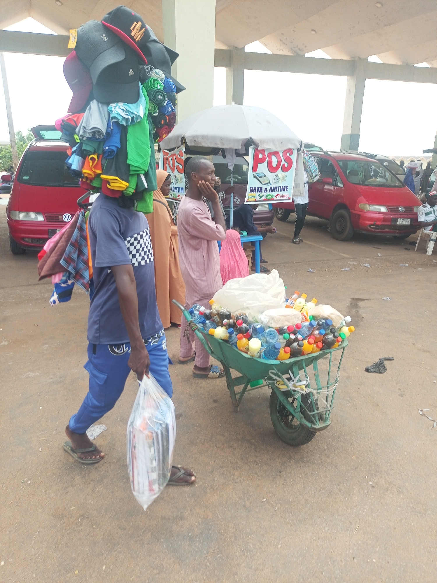 The image captures a bustling outdoor market or street scene in Gombe, Nigeria, under clear daylight. The ground is unpaved and dusty.
The central figure is a young man walking towards the left. He is wearing a blue short-sleeved t-shirt with a black and white checkerboard pattern on the chest, and blue cargo-style trousers. He has dark skin and is wearing flip-flops. On his head, he expertly balances a towering stack of merchandise, primarily baseball caps of various colors (black, red, grey) and folded t-shirts or clothing items in blue, green, yellow, and black. He carries a transparent plastic bag containing what appears to be folded items or newspapers in his right hand.
To the right of the walking man, a green metal wheelbarrow is laden with numerous plastic bottles containing colorful liquids (likely soft drinks and water), along with some white plastic bags containing snacks or food items. Another man, dressed in a light-colored traditional garment (agbada or similar), stands near the wheelbarrow, looking to the left. He has his hand resting on his chin.
In the background, under a white umbrella, a "POS" (Point of Sale) sign is prominently displayed. Below "POS", the text "DATA & AIRTIME" is visible, along with logos for telecommunication networks: MTN, GLO, and Airtel, indicating a service for mobile top-ups and data sales. Behind this stall, a red minivan (possibly a Volkswagen Sharan or similar) is parked with its rear door open. Another red minivan is visible further back on the right, and other people are scattered throughout the background, some sitting or standing. A large concrete overhang structure provides shade over parts of the scene. The overall impression is one of active street vending and commerce.