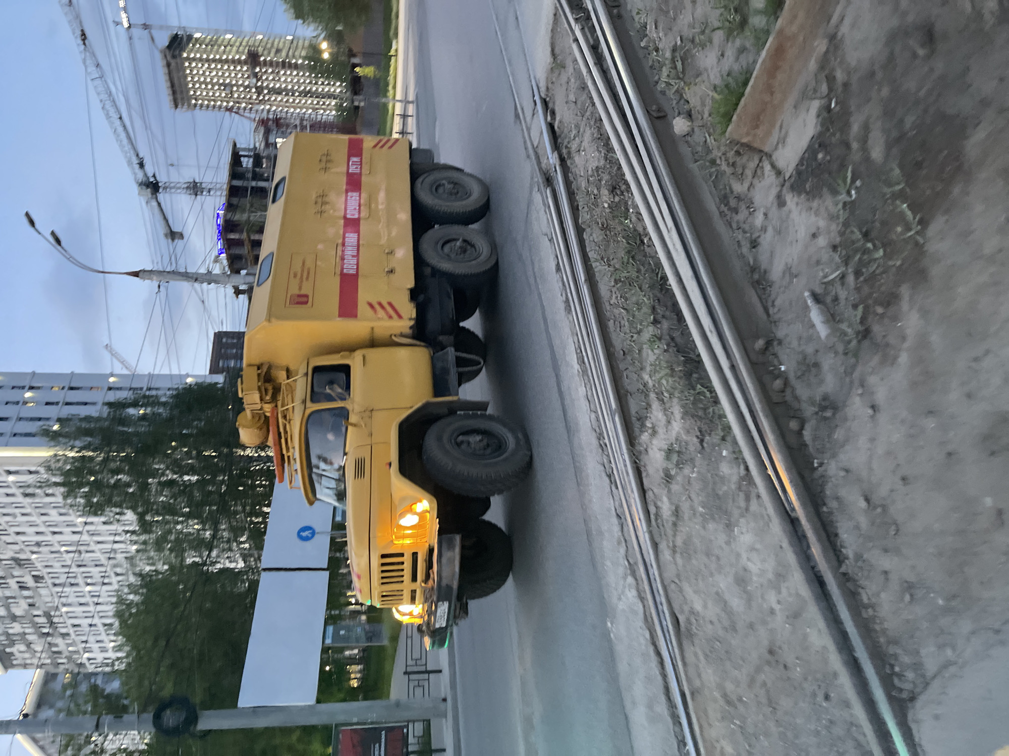 The image depicts a yellow maintenance truck driving on a road next to tram tracks in the city of Izhevsk, Russia. Construction is underway in the background, with cranes and building frameworks visible. The truck is equipped with multiple axles and appears to be a specialized vehicle. The sky is overcast, suggesting dusk or dawn. Pedestrian crossing signs are visible near the truck's front.