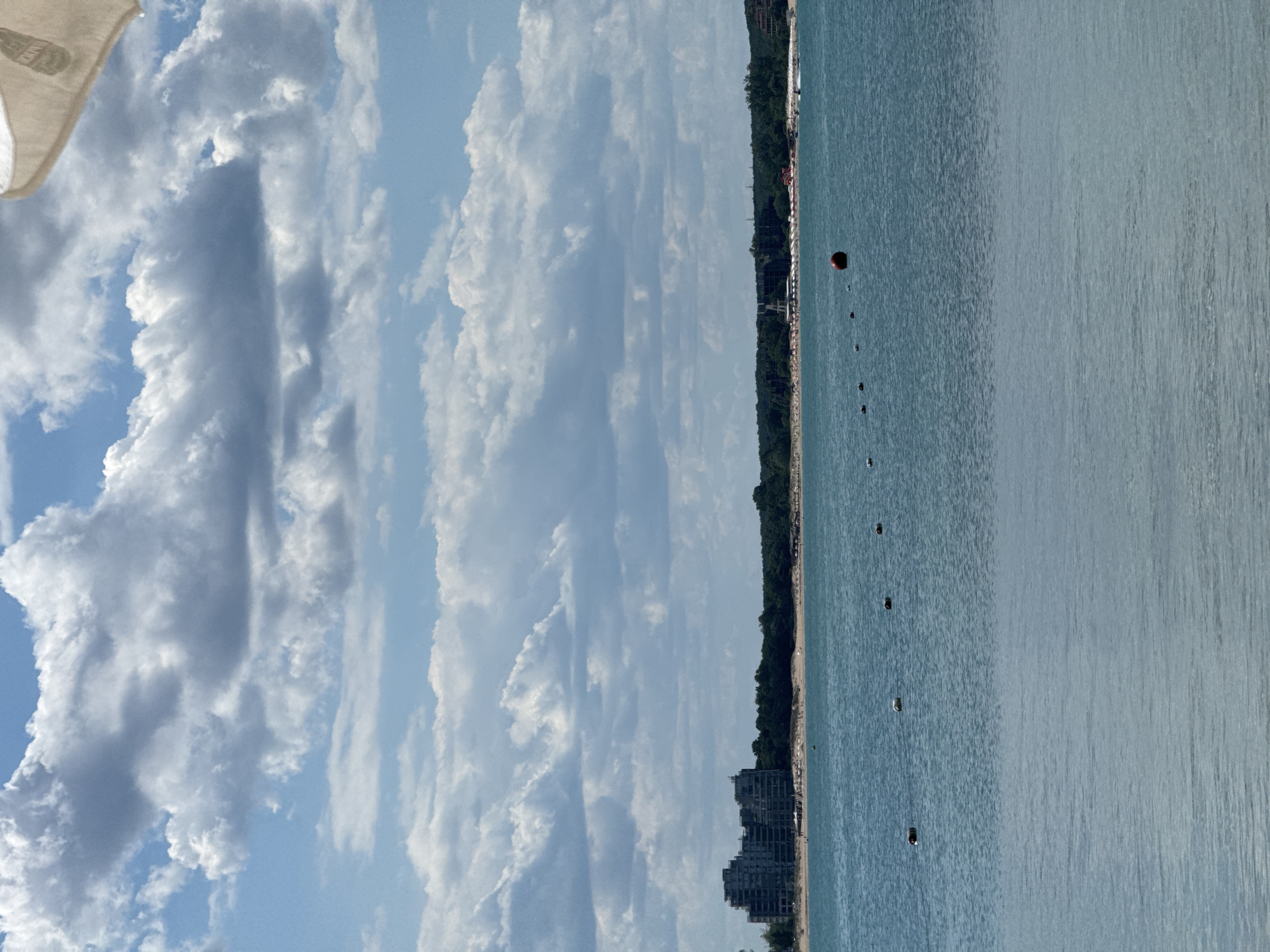 The image depicts a coastal scene in Kiten, Bulgaria, with a body of calm, blue water in the foreground and midground. Along the distant shoreline, there is a sandy beach lined with dense green trees. Several buildings, appearing to be hotels or apartment complexes, are visible behind the trees. The sky is filled with dramatic, textured white and gray clouds against a pale blue backdrop. A few small buoys are scattered across the water, and one larger red buoy is prominently positioned in the midground. The edge of a light-colored umbrella is visible in the upper right corner of the frame, suggesting a beach umbrella. The overall impression is a serene summer day at a seaside resort.