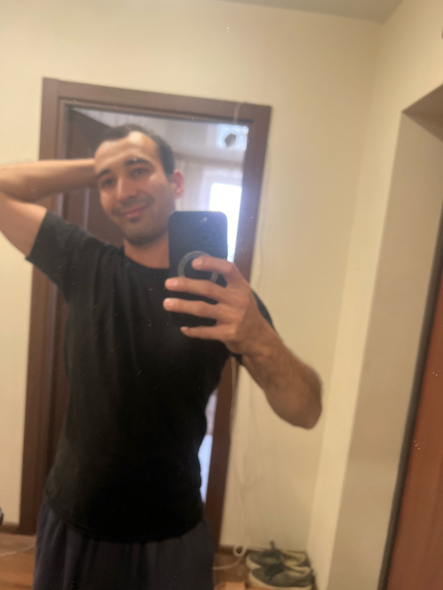 A medium shot shows a man posing for a mirror selfie in what appears to be a residential hallway in Kazan, Russia. The man has dark hair and is wearing a black t-shirt and dark blue pants. He is holding his right hand up to the back of his head, and his left hand is holding a smartphone, which is capturing the reflection. In the background, a doorway and a portion of a shoe rack with sneakers are visible. The overall lighting is dim, suggesting the photo was taken indoors.