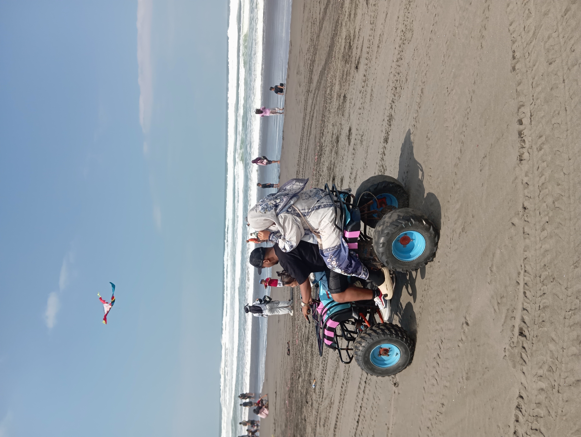 In Parangtritis, Indonesia, a man and a woman, both wearing headscarves, ride a colorful ATV on a sandy beach. The man, wearing a black t-shirt and shorts, drives while the woman, in a white and blue patterned outfit, sits behind him. A young child is seated in front of the man. In the background, waves crash onto the shore, and several people are visible, some standing and others sitting on the sand. The sky is clear with a few wispy clouds, and a colorful kite is flying high above. Tire tracks are visible in the sand, indicating the movement of the ATV and other vehicles.