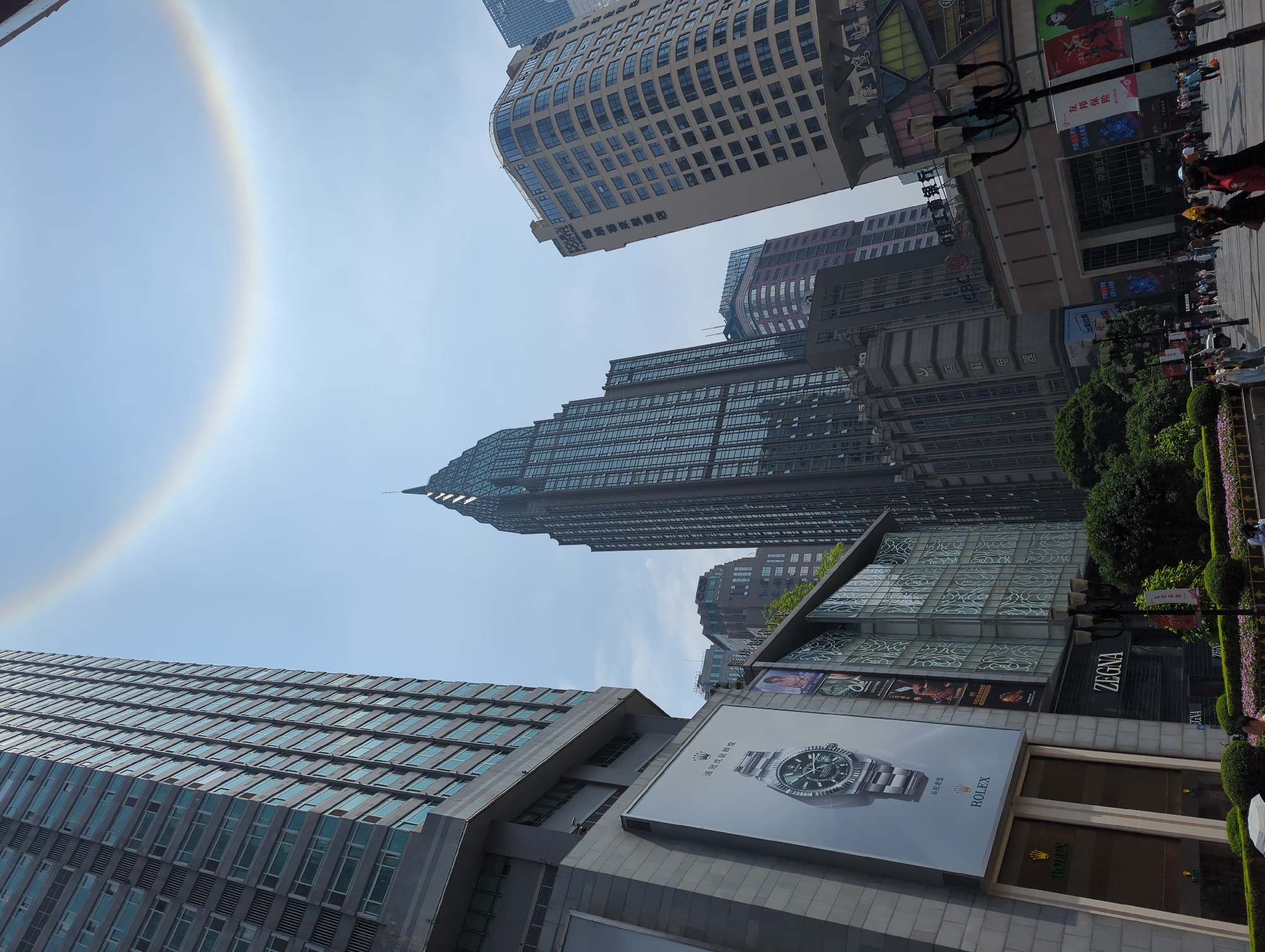 Here is a detailed description of the image:
**General Overview:**
The image captures a street scene in a densely built-up urban environment, possibly within the Yuzhong District of China. The viewpoint is from ground level, looking upwards at a cluster of tall buildings, street-level commercial areas, and the sky above.
**Environment and Architecture:**
* **Buildings:** The image features a variety of tall buildings, primarily skyscrapers with glass facades. The architectural styles vary, with some buildings having a sharp, pointed apex (like a spire), and others having more boxy, modern designs.
* **Commercial Area:** There are shop fronts with signs for brands such as "ROLEX," "ZEGNA," and "ICBC." These suggest a high-end retail or financial district.
* **Sky:** The sky is visible, and there is a visible optical phenomenon resembling a halo or rainbow arc in the upper-left corner of the image.
**Objects:**
* **Street Lamp:** A street lamp is situated on the right side of the image.
* **Advertisements:** Large advertisements or billboards for luxury watch brands (Rolex) are visible on the lower sections of the buildings.
**People:**
* **Crowd:** A group of people can be seen on the right side of the frame.
**Actions and Interactions:**
The scene suggests a busy urban environment. The people may be shopping or passing through the area.
**Specific Details:**
* The "ICBC" sign suggests the presence of the Industrial and Commercial Bank of China.
* The prominence of brands like Rolex and Zegna indicates a high-end commercial environment.
* Optical Phenomenon: The circular rainbow or halo-like effect in the sky is a noteworthy atmospheric phenomenon, possibly a solar halo.
**Location Hypothesis:**
Based on the architectural style and commercial brand presences, the image possibly depicts a prominent commercial district in the Yuzhong District, China.
