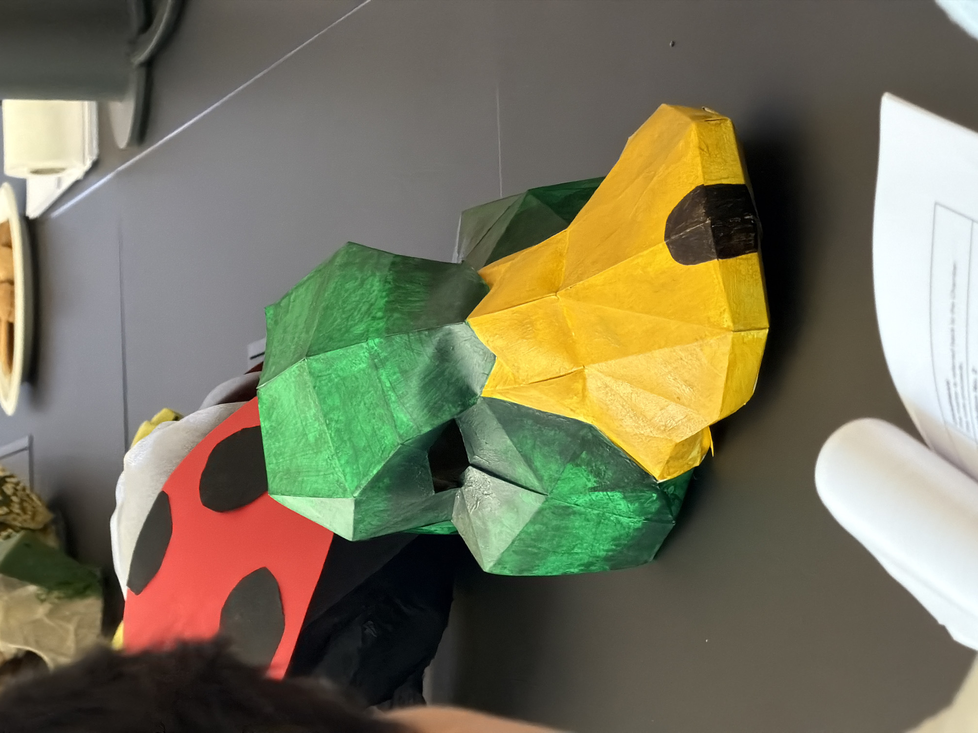 A close-up, high-angle shot captures a portion of a person's dark hair on the left, obscuring their face. In the center, a stylized, polygonal duck figurine is the main subject. The duck's head and beak are a bright yellow with a dark brown or black marking on the tip of its beak. Its body is comprised of faceted green segments. Behind the duck, a red and black spotted object, possibly a ladybug decoration, is partially visible. The objects are placed on a dark, matte surface, likely a table. In the background, out of focus, are some papers, a white roll of tape or paper towels, and a portion of a monitor or desk lamp. The scene is indoors, and the lighting appears to be artificial. The image does not contain any information or visual cues that would suggest a specific location in Milan, Italy.