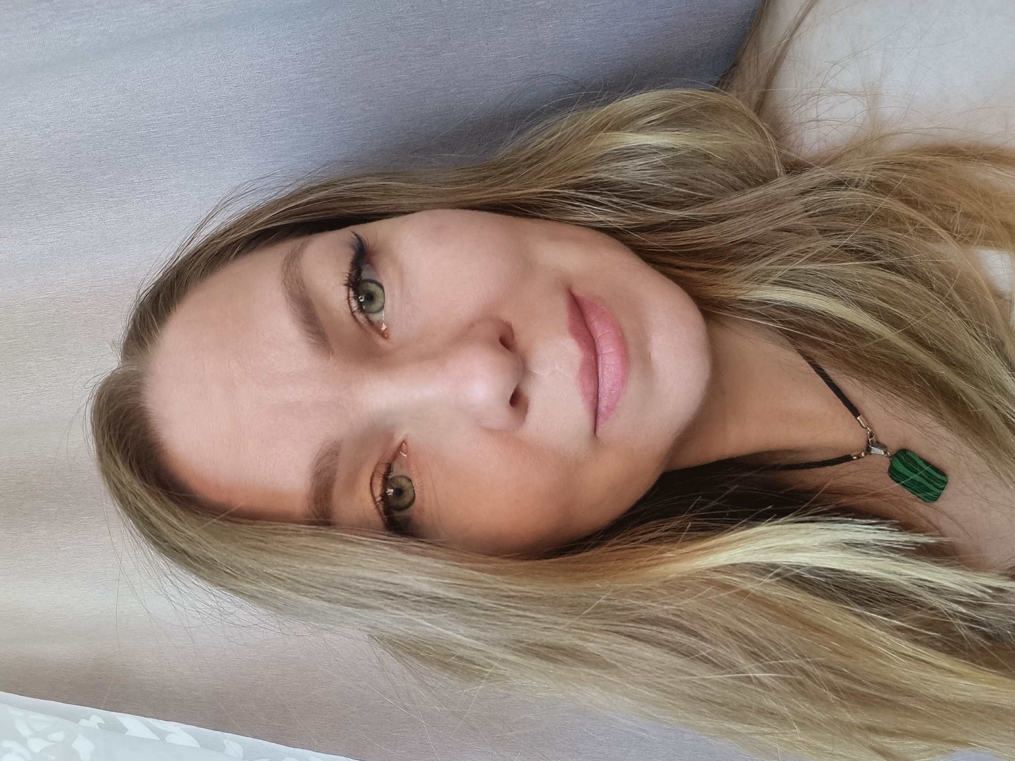 A close-up, rotated image depicts a Caucasian woman with light blonde hair and green eyes. Her skin is fair, and she is wearing light makeup, including eyeliner and lip color. A necklace with a green, rectangular pendant is visible around her neck. The background is a light grey textured surface, possibly fabric. The image was taken indoors. The location, Chaikovsky, Russia, cannot be verified from the image.