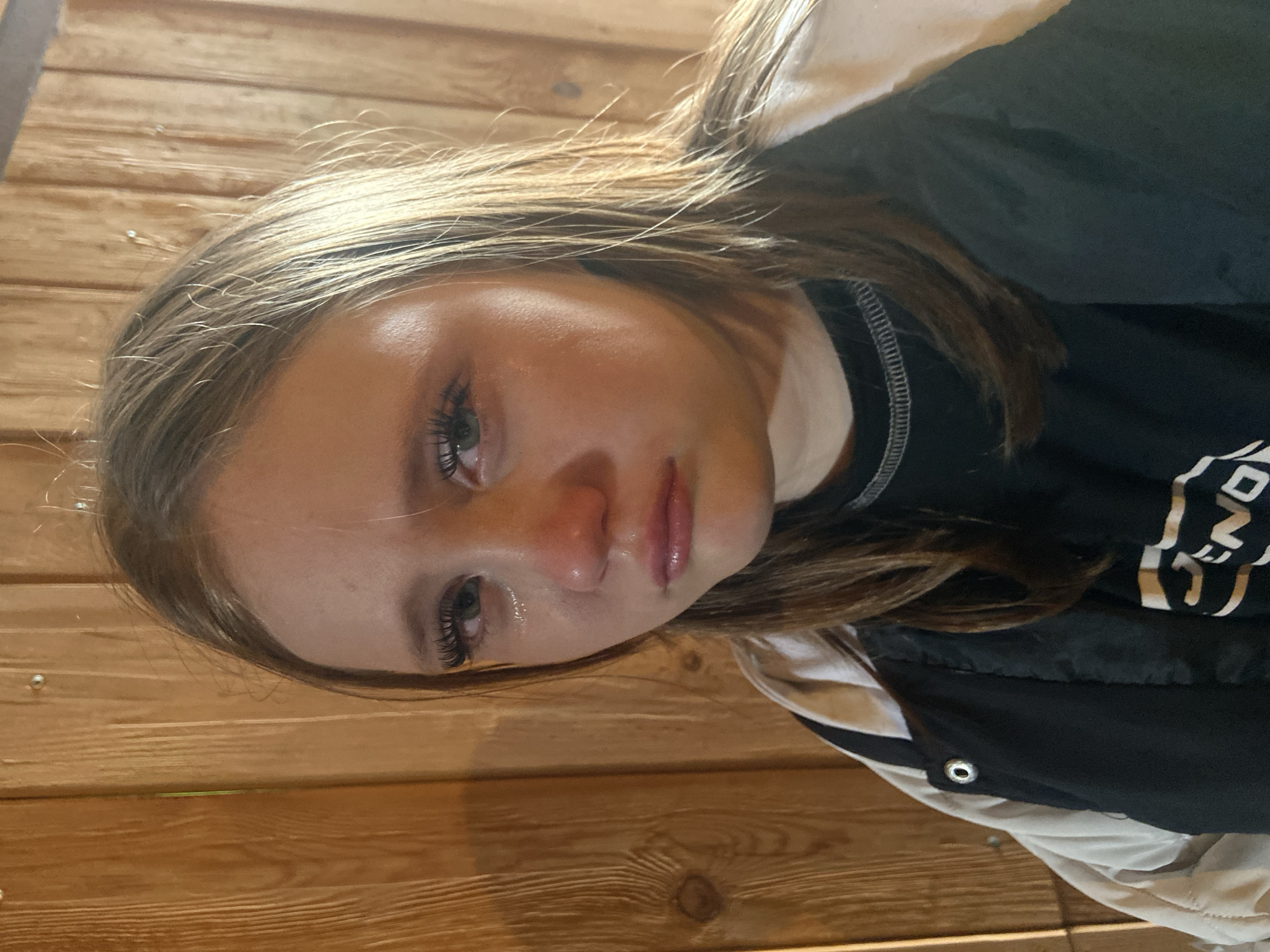 A close-up, indoor shot shows a young female with light blonde/brown hair and fair skin. She appears to be lying down, her head slightly angled. Her eyes are open, possibly watery. She is wearing a black shirt with white graphic elements and a dark jacket with a white stripe and what seems to be a silver grommet. The background is a wood paneling wall. There's no information available about the city of Chaikovsky, Russia, or a location in that city based on the image.