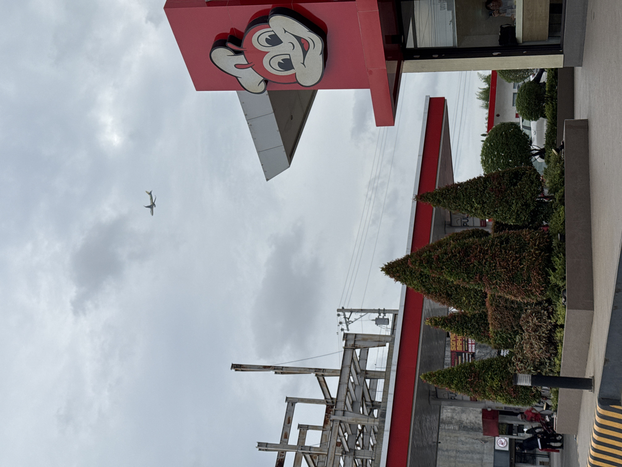 This outdoor photograph, taken in Davao City, Philippines, depicts a bustling commercial environment under an overcast sky. The most prominent feature is a large, red sign adorned with the iconic smiling face of the Jollibee mascot, angled towards the viewer from the upper left portion of the frame. In the cloudy sky above, a small airplane is visible in flight.
The scene includes various urban elements. To the right, several manicured, conical evergreen shrubs with a mix of green and reddish foliage line a paved area, which features a black and yellow striped curb in the foreground. A black street lamp or bollard is positioned among these plants. In the background, a white vehicle and a motorcycle with a person wearing a red shirt are partially visible. A red circular sign, likely indicating "No Stopping" or "No Entry," is also present.
Part of a building with red and gray facades is visible, with a window reflecting what appears to be a person wearing glasses. Below the Jollibee sign and towards the lower left, an unfinished metal framework structure, possibly for a building extension or canopy, stands tall. Utility poles and power lines stretch across the sky near this structure. A partially obscured white sign, visible through the plants, displays text that includes "SIGNAGE MAKER" and several phone numbers. The weather is clearly overcast, with a bright but cloudy sky suggesting daytime.