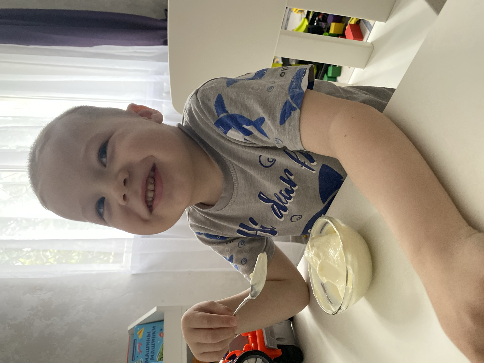 A joyful young boy is captured in a brightly lit indoor setting, likely in Sochi, Russia, as he enjoys a snack. The image, taken from a slightly elevated perspective, shows the boy looking directly at the camera with a wide, radiant smile, indicating happiness and contentment. He has short, light-colored hair and is wearing a gray t-shirt adorned with blue shark or whale patterns and some decorative script.
He is seated at a simple white table, holding a silver spoon with a creamy, light-colored substance, possibly yogurt or pudding, which he has taken from a small, clear glass bowl placed in front of him. The activity suggests a casual mealtime or snack break.
The background reveals a large window with sheer white curtains, allowing natural daylight to illuminate the scene and offering a glimpse of lush greenery outside. Below the window, a white shelf holds a blue and red book with the Cyrillic text "МАШИНЫ МАШИНКИ" (Cars, Toy Cars). Further back, colorful toys, possibly building blocks, are visible on another shelf, and a red and black toy truck can be partially seen on a surface to the lower right. The bright lighting confirms it is daytime.