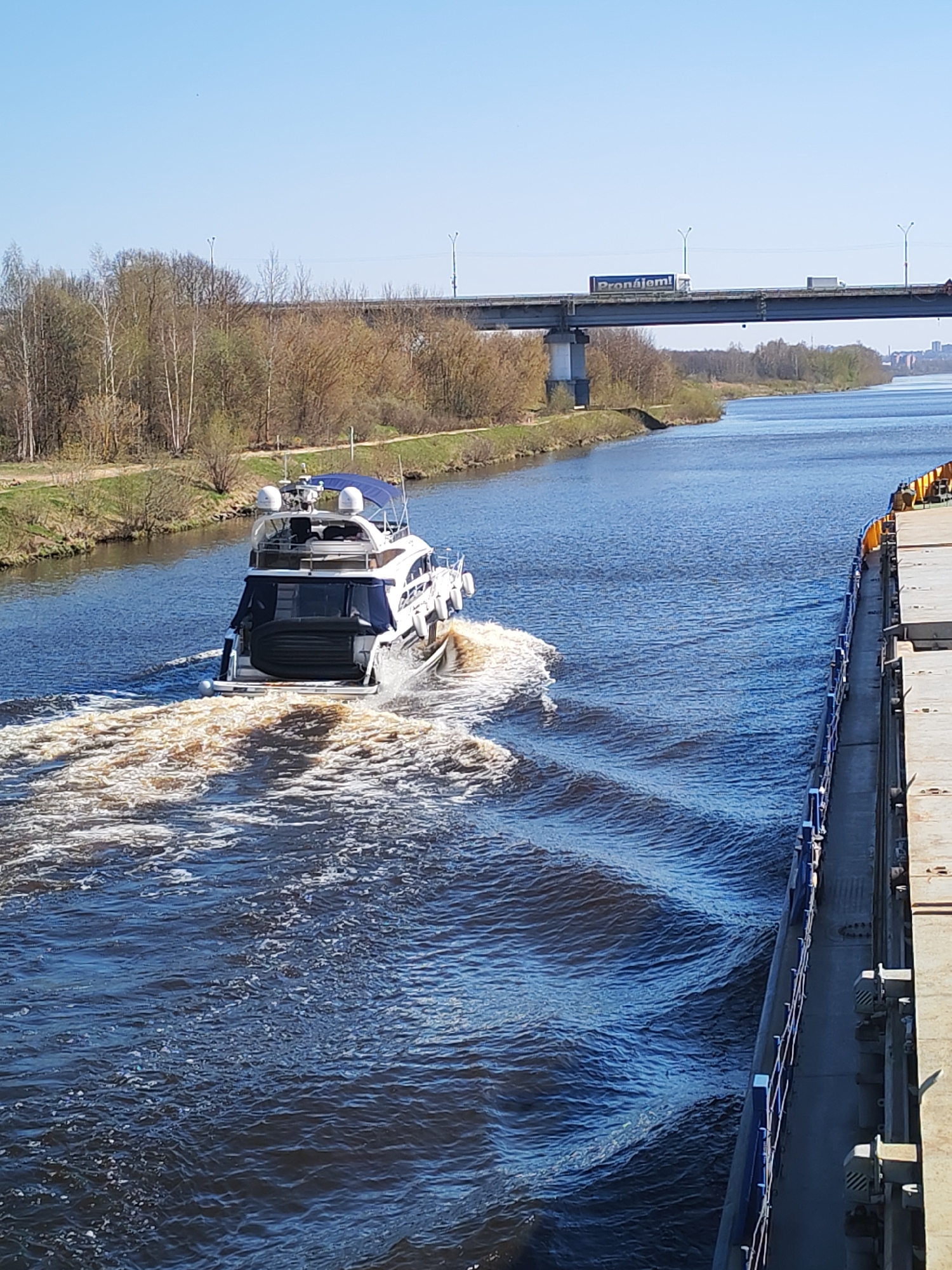 A medium shot depicts a motor yacht traveling along a canal, generating a foamy wake. To the right of the yacht, the side of a barge or larger vessel is visible. A bridge spans the canal in the background with a blue sign, partially visible, reading "Pronájom!" in white letters. The canal banks feature a line of trees, and the sky is clear and blue. The image, taken from a high vantage point, suggests the location is the city of Druzhba 6, Russia, identifiable by the bridge signage.