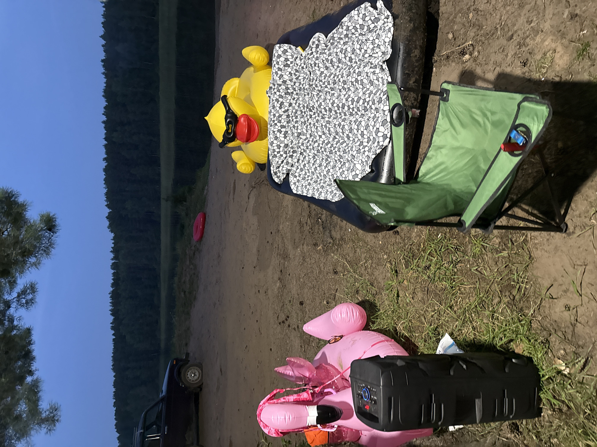 The image depicts an outdoor scene by a body of water, possibly a lake or river, with a dense forest line in the background. The time appears to be dusk or twilight, with a muted blue sky. In the foreground, on a sandy and grassy area, are several objects. To the left, a large pink inflatable flamingo float is partially deflated and positioned near a black portable speaker. To the right, a bright yellow inflatable duck with black sunglasses and a red beak is resting on a dark inflatable raft covered with a black and white patterned blanket. A green folding camping chair is placed in front of the raft, with a red object and a blue object visible in its cup holder. In the middle distance, a red inflatable ring or float is visible on the ground. A dark-colored SUV is parked on the left side of the image, facing away from the water. The overall impression is a relaxed, recreational setting, potentially a campsite or picnic area by the water. The specific location in Chaykovsky, Russia, is not explicitly identifiable from the visual elements alone, but the environment is consistent with natural landscapes found in the region.