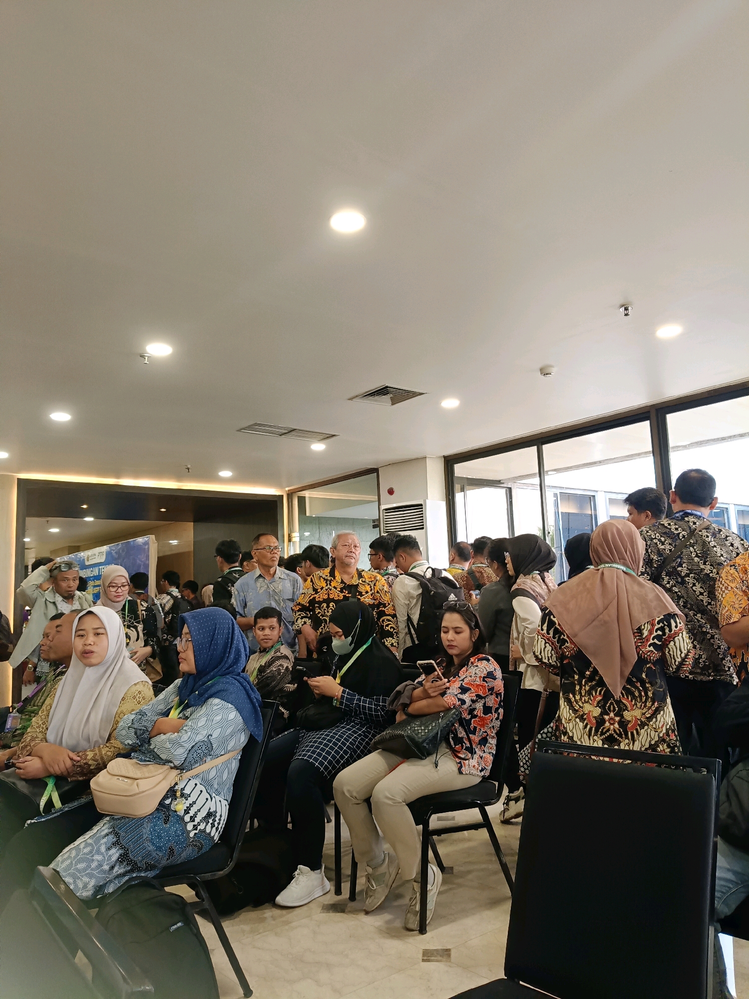 A medium shot shows a group of people gathered indoors, possibly at an event or conference. The lighting is bright, with recessed lights visible in the ceiling. A banner with Indonesian text is partially visible on the left. Several individuals are seated in chairs, while others stand. Many are wearing batik-patterned clothing, suggesting a cultural or formal occasion. Some people are looking at their phones, while others are engaged in conversation or observing their surroundings. The environment appears to be a modern building with large windows. The overall scene depicts a populated indoor gathering.