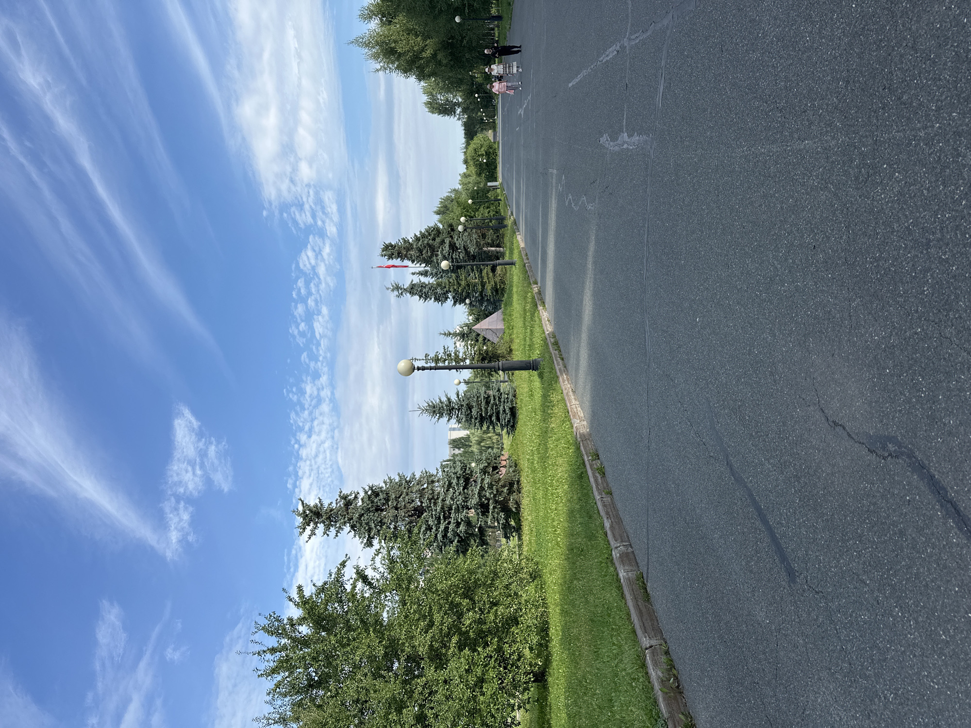 The image depicts a wide asphalt path in a park-like setting with lush green grass and trees lining the left side. The path extends into the distance, where several people are walking. On the left, there are tall evergreen trees and a deciduous tree. Lampposts with spherical lights are visible along the path. In the background, there is a large pyramid-shaped structure with a red flag on a flagpole to its left. Buildings are visible in the far distance. The sky is blue with wispy white clouds. The scene appears to be a public park, possibly in Kazan, Russia, given the context provided in the prompt. The image captures a serene outdoor environment on what appears to be a pleasant day.