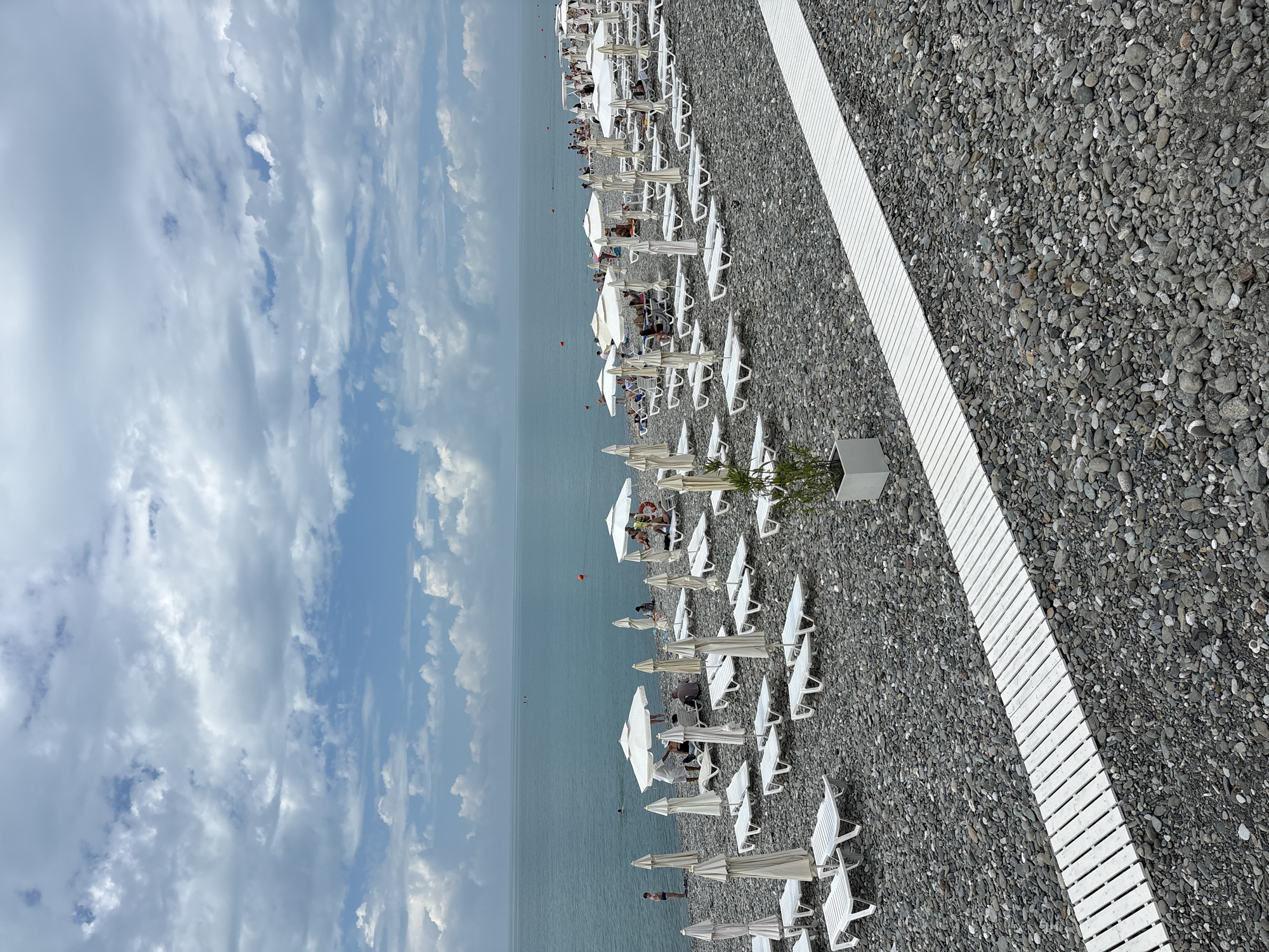 This is a medium-shot, eye-level, outdoor photograph of a pebble beach in Sochi, Russia. The beach is lined with rows of white lounge chairs and umbrellas. A white wooden walkway runs across the foreground of the image, diagonally from the bottom left to the top right. The sea is a deep blue and stretches to the horizon, where it meets a cloudy, overcast sky with patches of blue. Several people are visible on the beach, some sitting on lounge chairs and others swimming in the sea. There are also a few buoys in the water. In the foreground, to the right, a potted plant sits next to the walkway. The overall scene depicts a popular tourist destination during daylight hours.