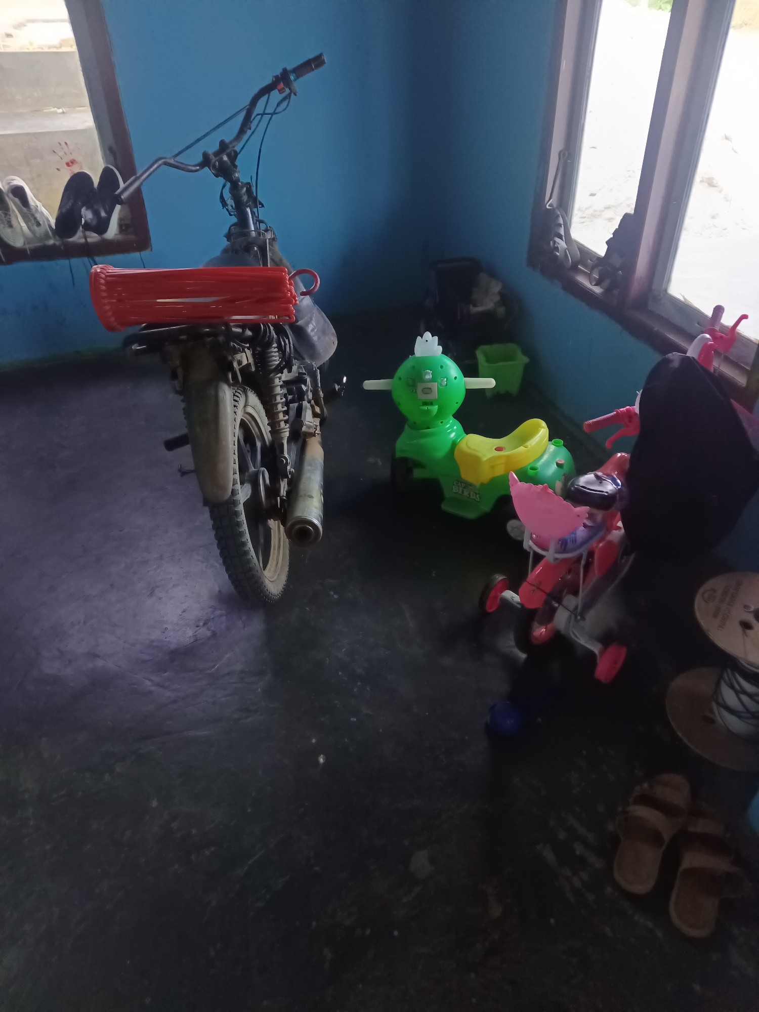 The image depicts an indoor scene with a motorcycle and several toys. The motorcycle is positioned on the left side of the frame, facing away from the viewer. It is a dirt bike with a rear rack holding a red rectangular object, possibly a storage container or a bundle of items. The motorcycle's rear wheel and exhaust pipe are clearly visible.
To the right of the motorcycle, there are several toys. A bright green ride-on toy with a yellow seat is present, and next to it is a pink tricycle-like toy with a black seat and red handlebars. A black bag is draped over the pink toy. In the background, there is a window with light streaming through, and a blue wall. On a shelf near the window, a pair of white and black shoes are visible. The floor is dark and appears to be concrete.
The setting is likely a residential space within the city of Terbanggi Marga, Indonesia. The presence of both a motorcycle and children's toys suggests a family home. The overall impression is one of a casual, lived-in space.