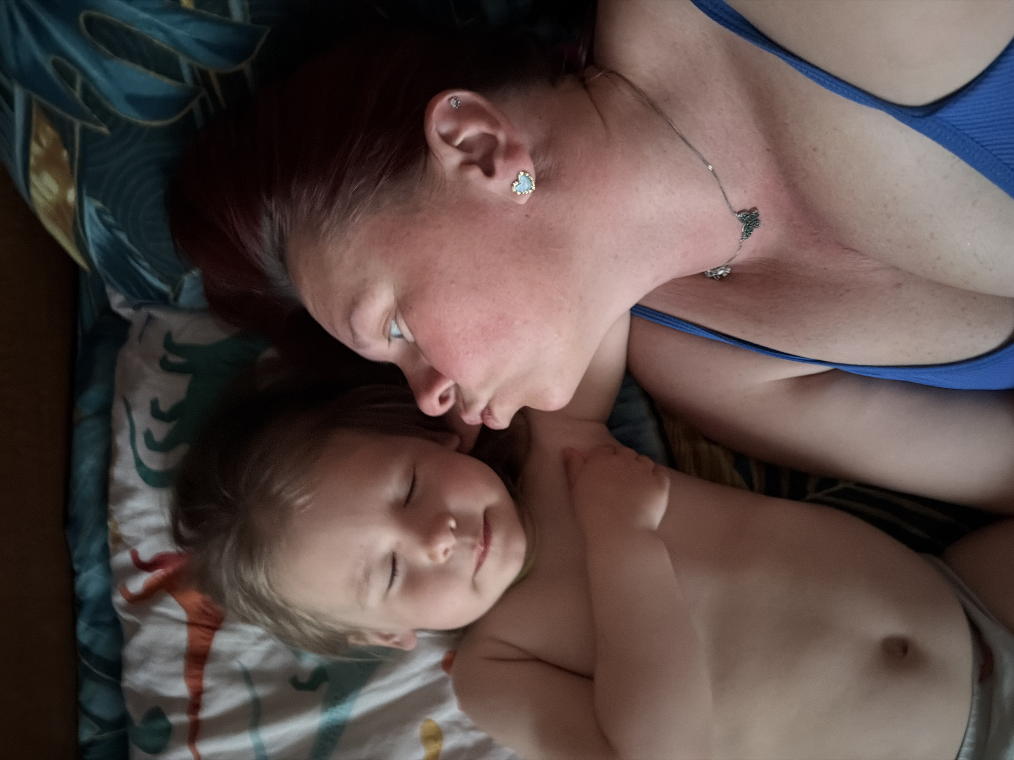 A top-down, medium shot depicts a woman and child lying on a patterned sheet. The woman has red hair, fair skin, and wears a blue top. She also has a necklace with a butterfly-shaped pendant and earrings, one with a heart shape and the other a small stud. The child is mostly unclothed, except for a diaper, with blonde hair, fair skin, and closed eyes. The woman appears to be nuzzling or kissing the child's forehead. The location is unknown and the city of Chaikovsky, Russia cannot be confirmed from the image.