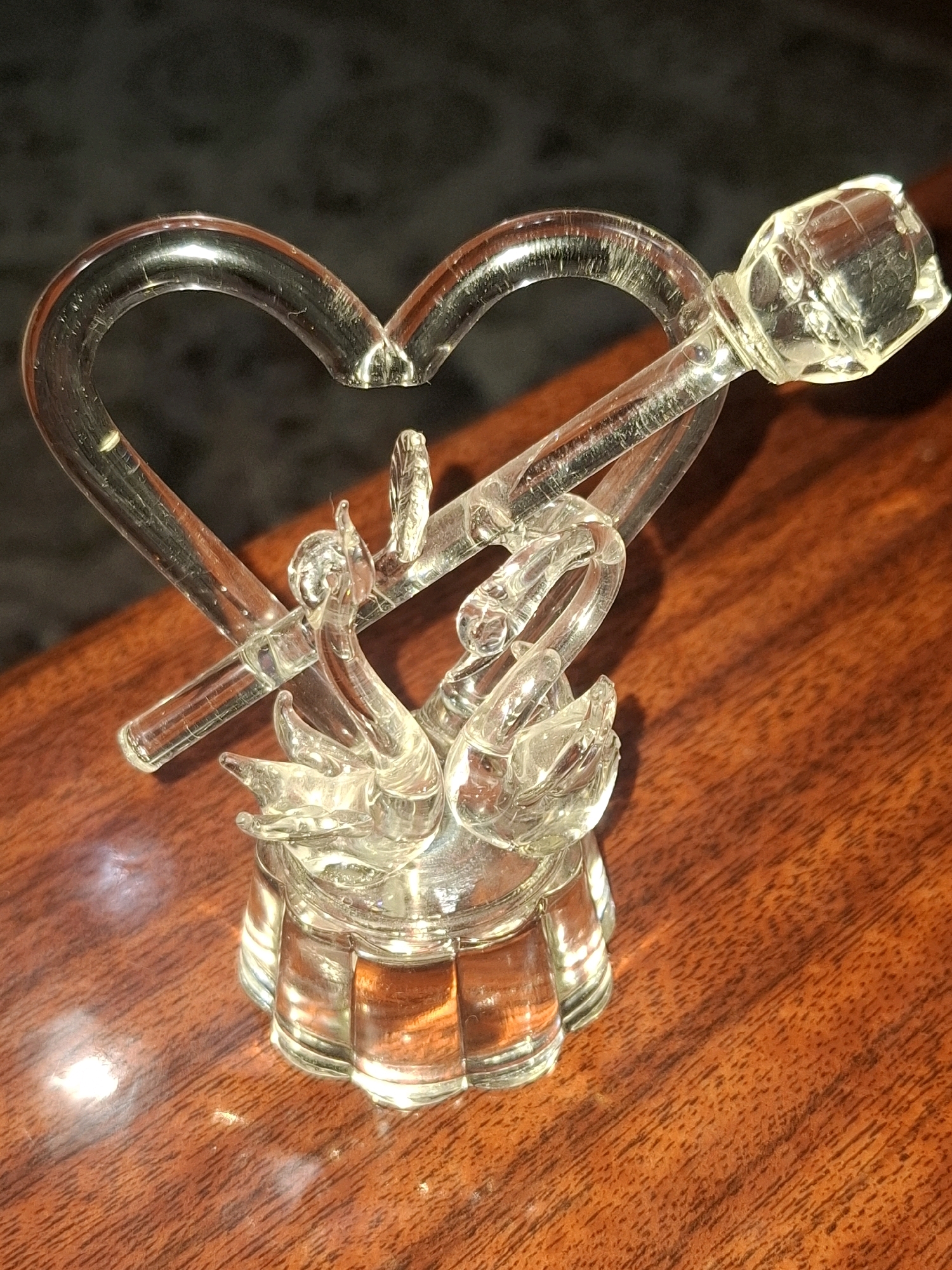 A clear glass figurine depicting two swans forming a heart shape with a rose at the end of a stem, rests on a polished wooden surface. The figurine is illuminated from above, casting shadows on the dark background behind it. The wood grain of the surface is clearly visible. No people or animals are present in the image. The scene appears to be indoors, possibly on a table or desk. The location of Almaty, Kazakhstan is not discernible from the image itself.