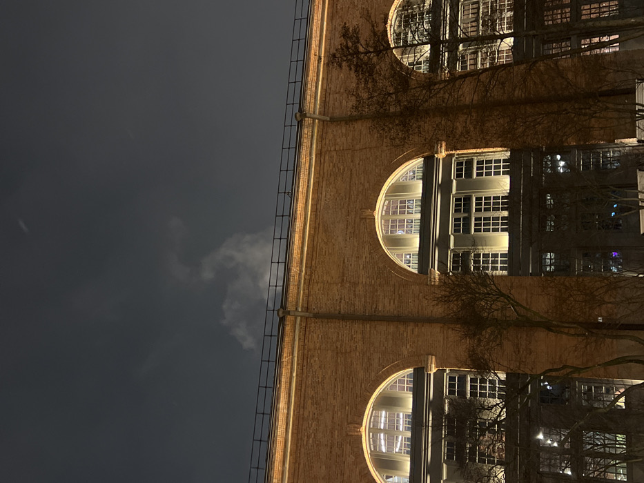 A tall brick building with rounded windows is shown against a dark sky. The windows are illuminated, and the building's surface appears to glow with a warm light. A ladder runs vertically up the building's side. Bare tree branches are visible in front of the building, partially obscuring the view. There are faint clouds in the sky. The perspective is looking up at the building from a low angle.