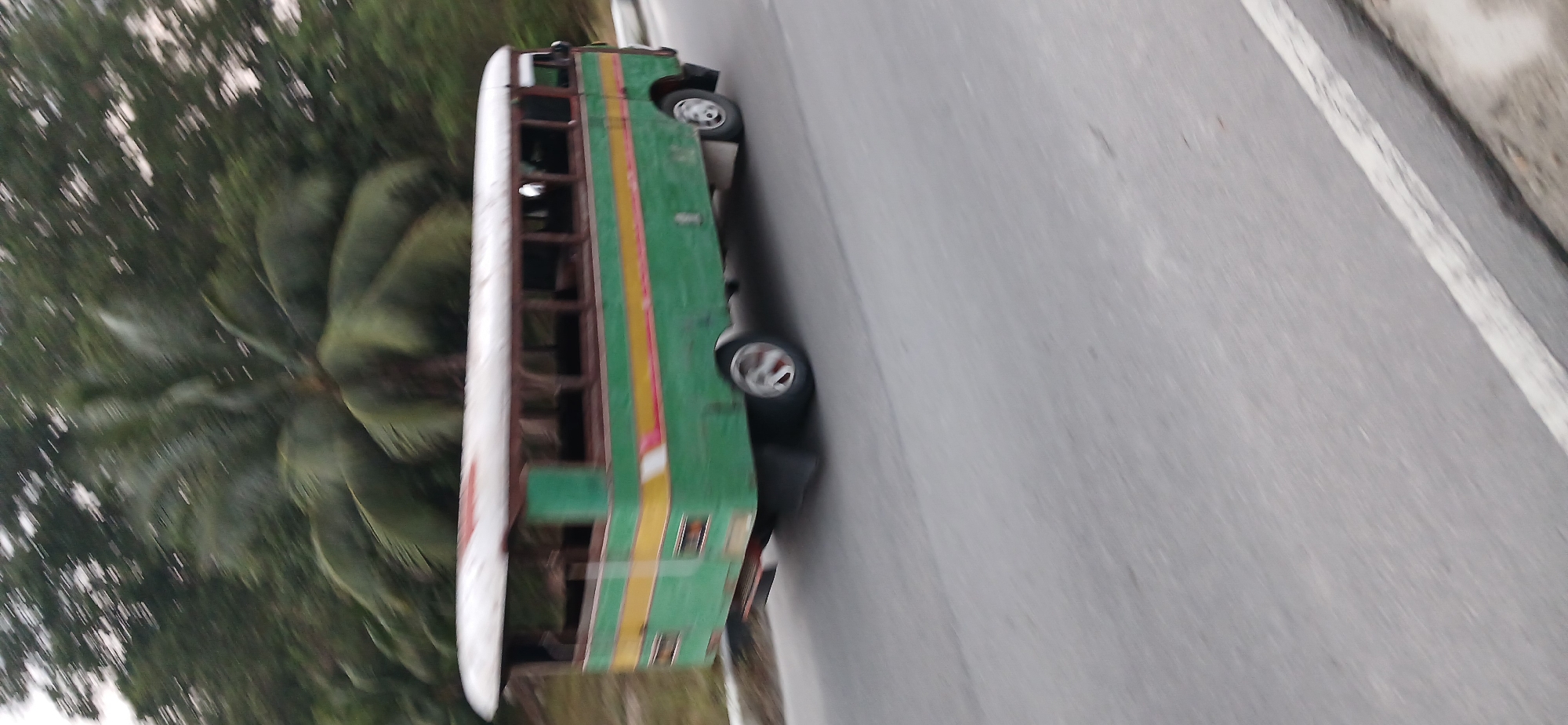 An old, green and yellow bus with a white roof is driving on a paved road, likely in the city of Tanjung Balai Karimun, Indonesia, as indicated by the hint. The bus is open-sided with visible seating. The background is blurred due to motion, but appears to be lush greenery with palm trees. The road surface shows a white line marking the edge. The image is captured in daylight.