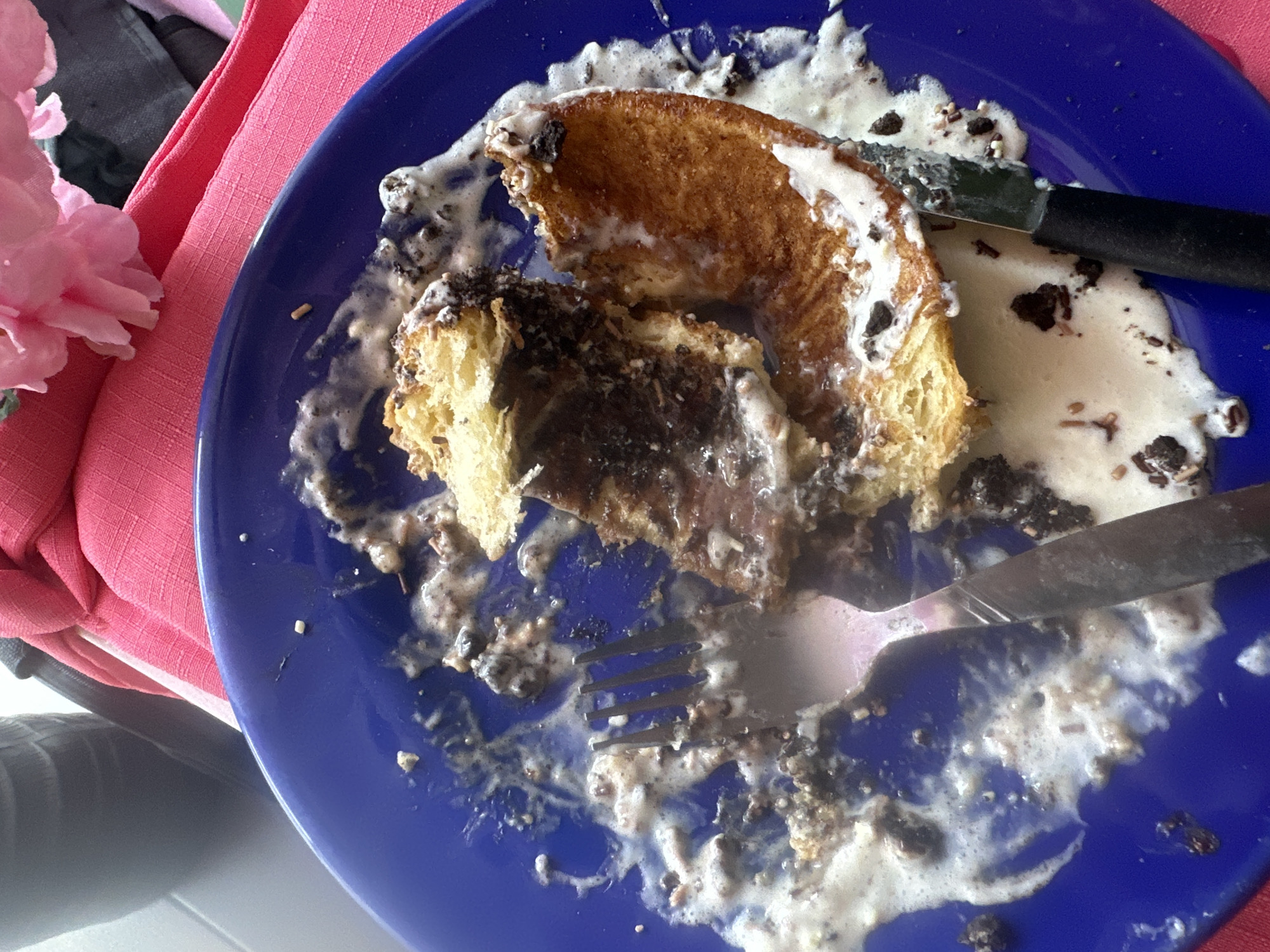 A partially eaten dessert is on a blue plate. The dessert consists of a pastry, possibly a type of bread or cake, with a dark, likely chocolate, filling. It is covered with a white sauce and dark sprinkles. A fork and knife are resting on the plate, indicating that the dessert was being eaten. The scene includes a pink cloth napkin and a pink flower on the side. The picture appears to have been taken indoors. There is no explicit information identifying that the photo was taken in San Andrés Cholula, Mexico.