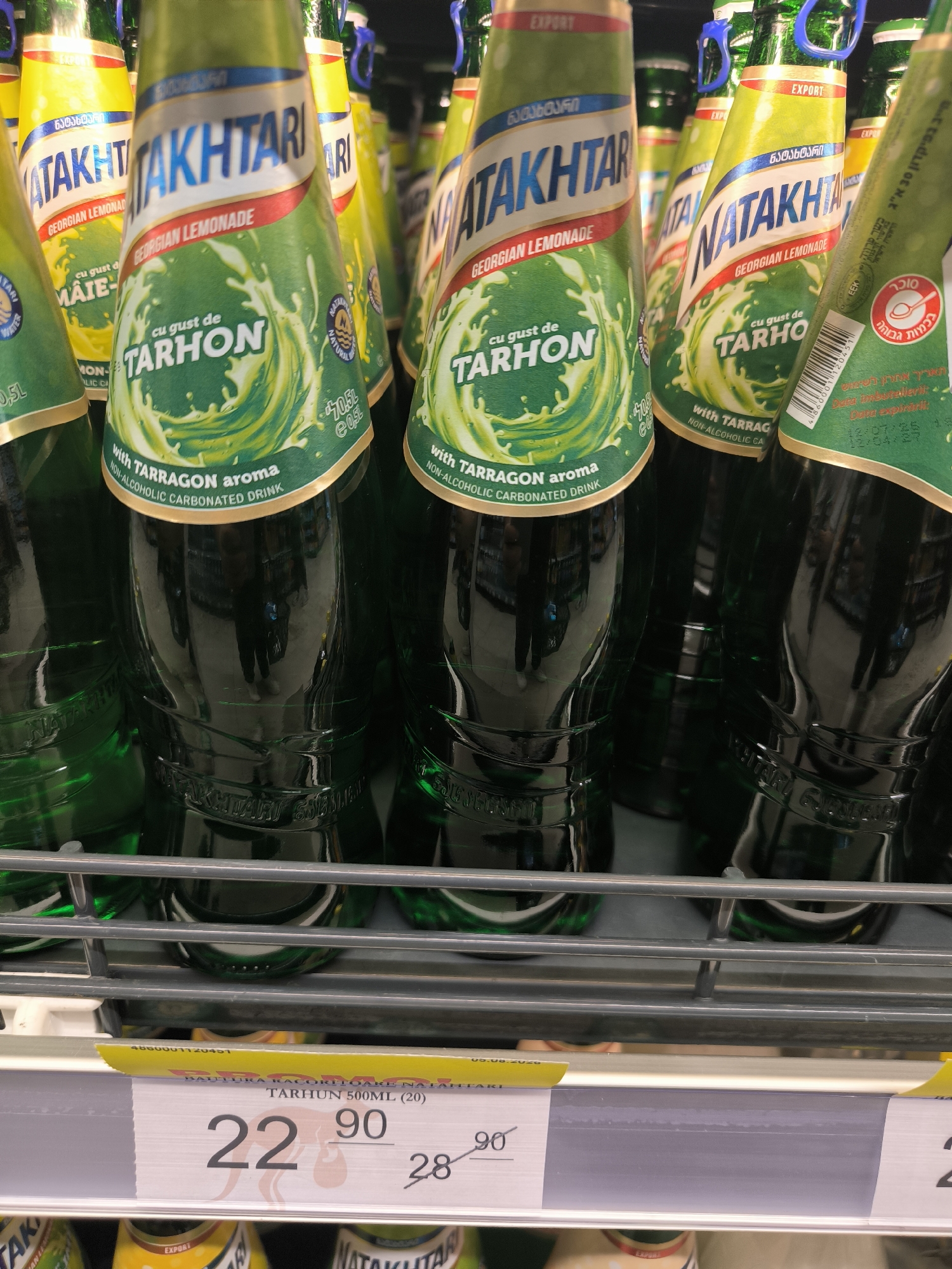 The image shows a retail shelf displaying multiple green glass bottles of "Natakhtari" brand Georgian Lemonade. The bottles are arranged closely together, with their labels prominently visible. The labels feature the brand name "Natakhtari" in large blue lettering, along with text indicating "Georgian Lemonade" and "cu gust de TARHON" (with the taste of Tarragon). Some bottles also have an "EXPORT" label at the top. In the foreground, a price tag is visible, showing a price of "22.90" and "28" with a slash through it, suggesting a sale or discount. The background of the image is blurred, but appears to be a retail store environment with other products and shelves. The bottles are reflections of the environment, including what appears to be a person in the background wearing dark trousers and light shoes. The image appears to have been taken at a supermarket in Chișinău, Moldova, based on the presence of a price tag with Cyrillic text and a price format common in the region.