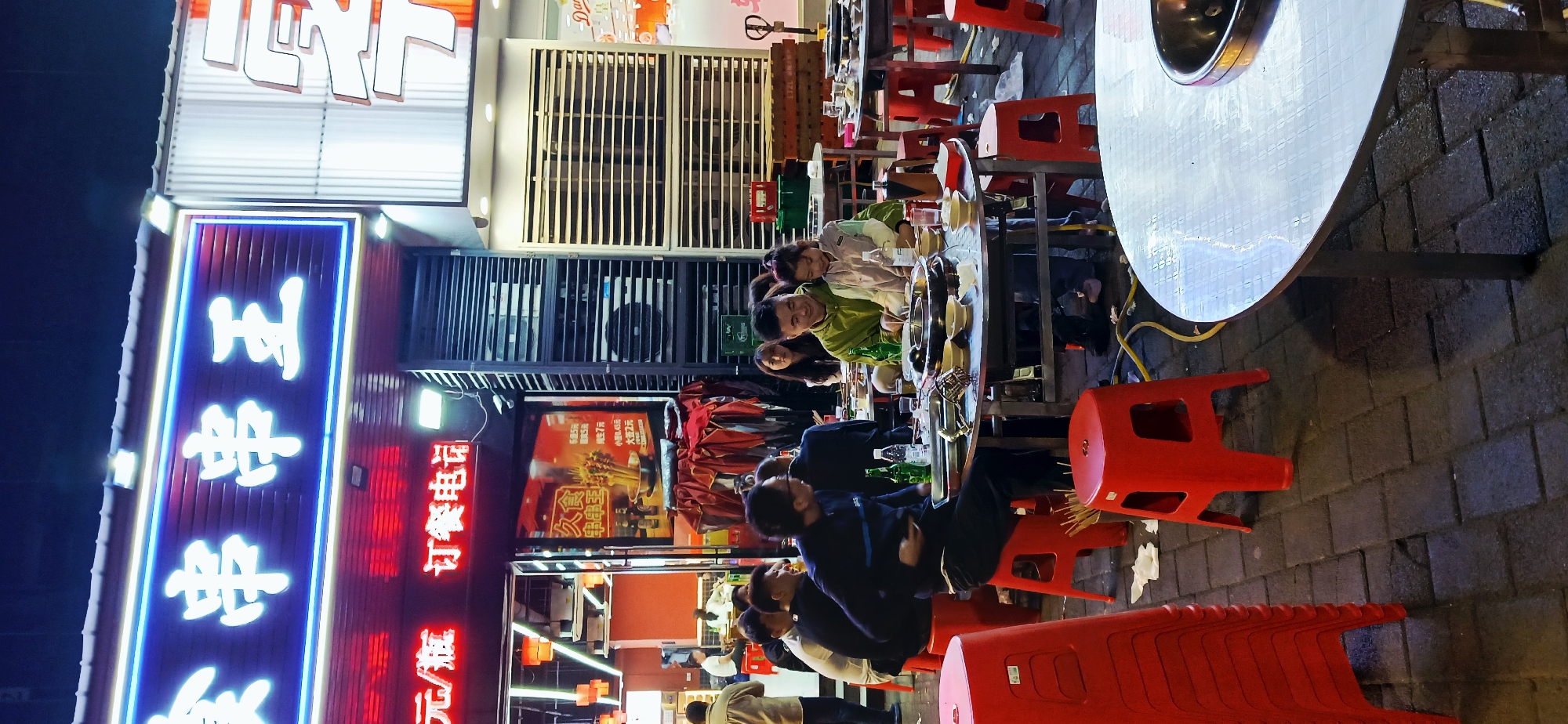 Here is a concise description of the image:
This is a night scene captured in what appears to be a street restaurant in the Jiulongpo District, China. Several people are sitting around a table enjoying a meal. The setting includes red plastic stools and tables prepared for hot pot. Neon signage with Chinese characters, including "串串王 (chuàn chuàn wáng)", indicates the name of the restaurant. The OCR text on the signs lists prices and ordering information. The setting suggests an outdoor dining area, possibly on a sidewalk.