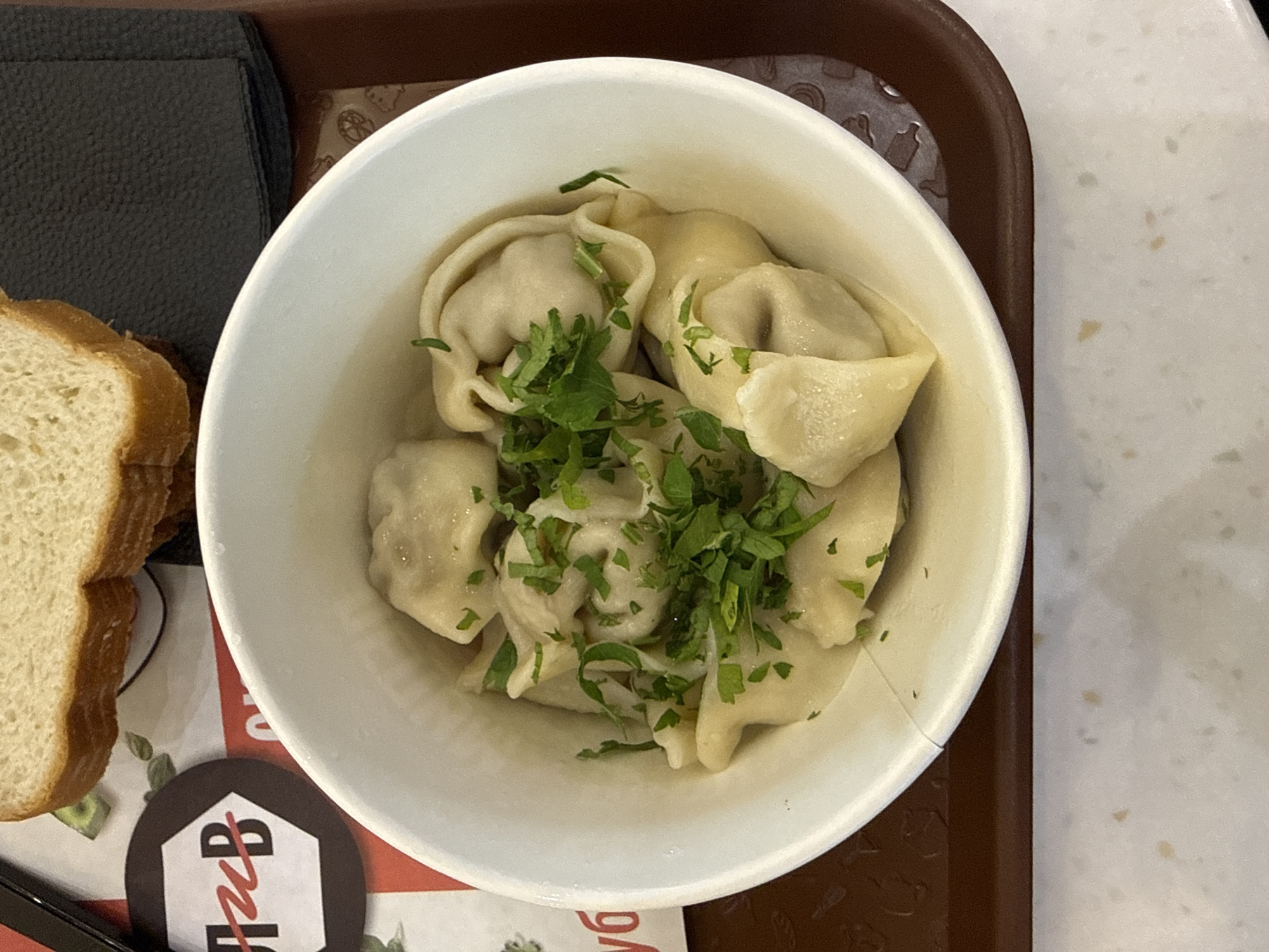 A close-up, overhead shot shows a white bowl filled with pelmeni, a type of Russian dumpling, topped with chopped fresh parsley. The bowl is placed on a brown tray. In the background, a slice of white bread rests on a paper placemat. To the left of the bowl, a circular logo with a hexagonal outline contains the Cyrillic letters "П и В" in red and white. The scene appears to be in a food establishment in Moscow, Russia, given the Cyrillic text. The lighting is bright, casting minimal shadows, suggesting an indoor setting with overhead illumination.