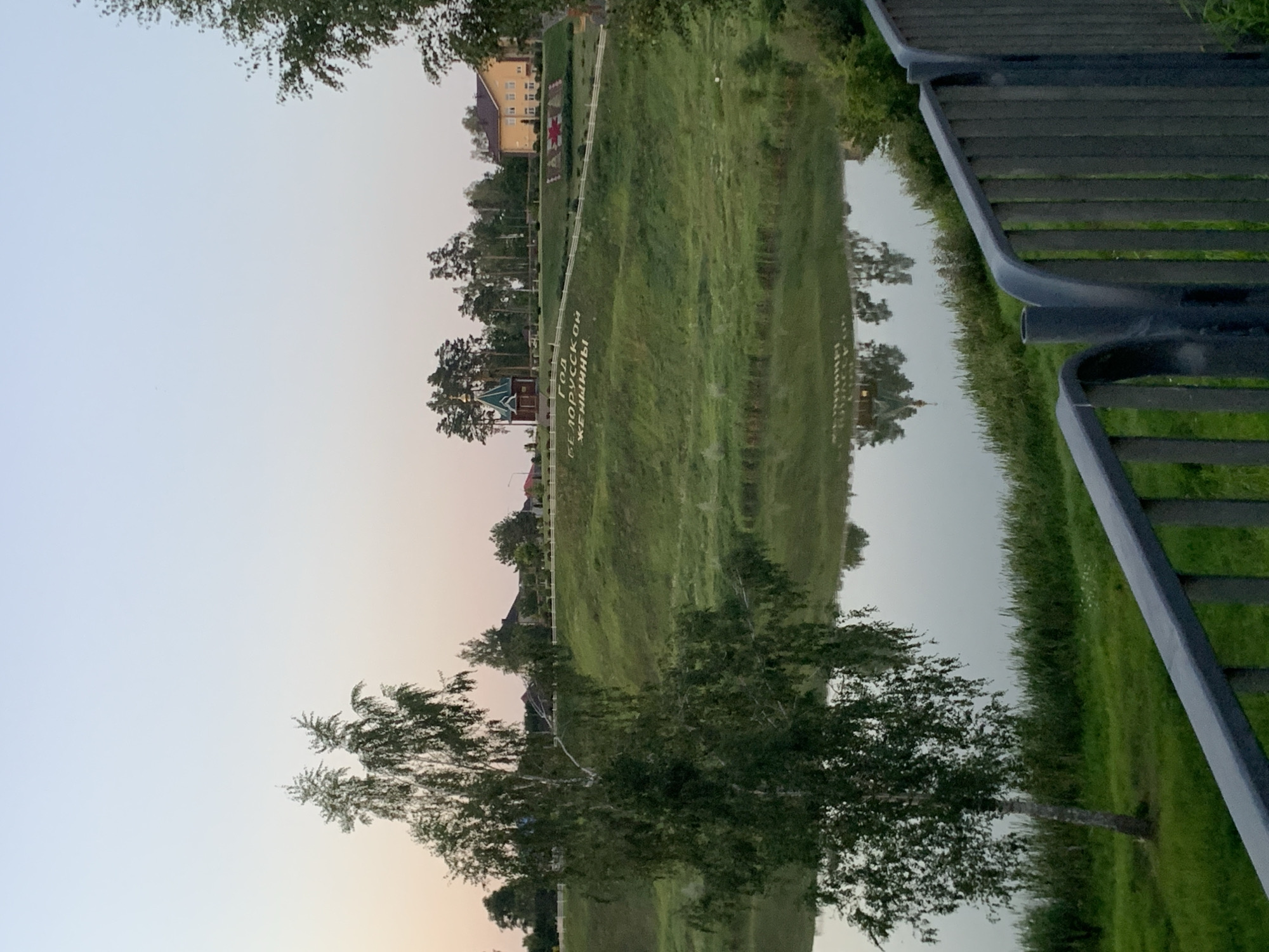 The image depicts a tranquil scene by a body of water, with a grassy hillside in the background. Inscriptions are visible on the hillside, which appear to be in Cyrillic script. A small, red and green-roofed building, possibly a chapel or church, is situated on the hillside, along with a yellow building and several trees. The water reflects the structures and trees on the shore, creating a mirrored image. A metal railing runs along the bottom right of the frame, suggesting the viewpoint is from a bridge or elevated pathway. The sky is a pale, soft hue, indicative of either dawn or dusk. The overall impression is one of peaceful, rural landscape with some man-made elements. The inscription on the hillside reads "ГОД БЕЛОРУССКОЙ ЖЕНЩИНЫ", which translates to "Year of the Belarusian Woman".