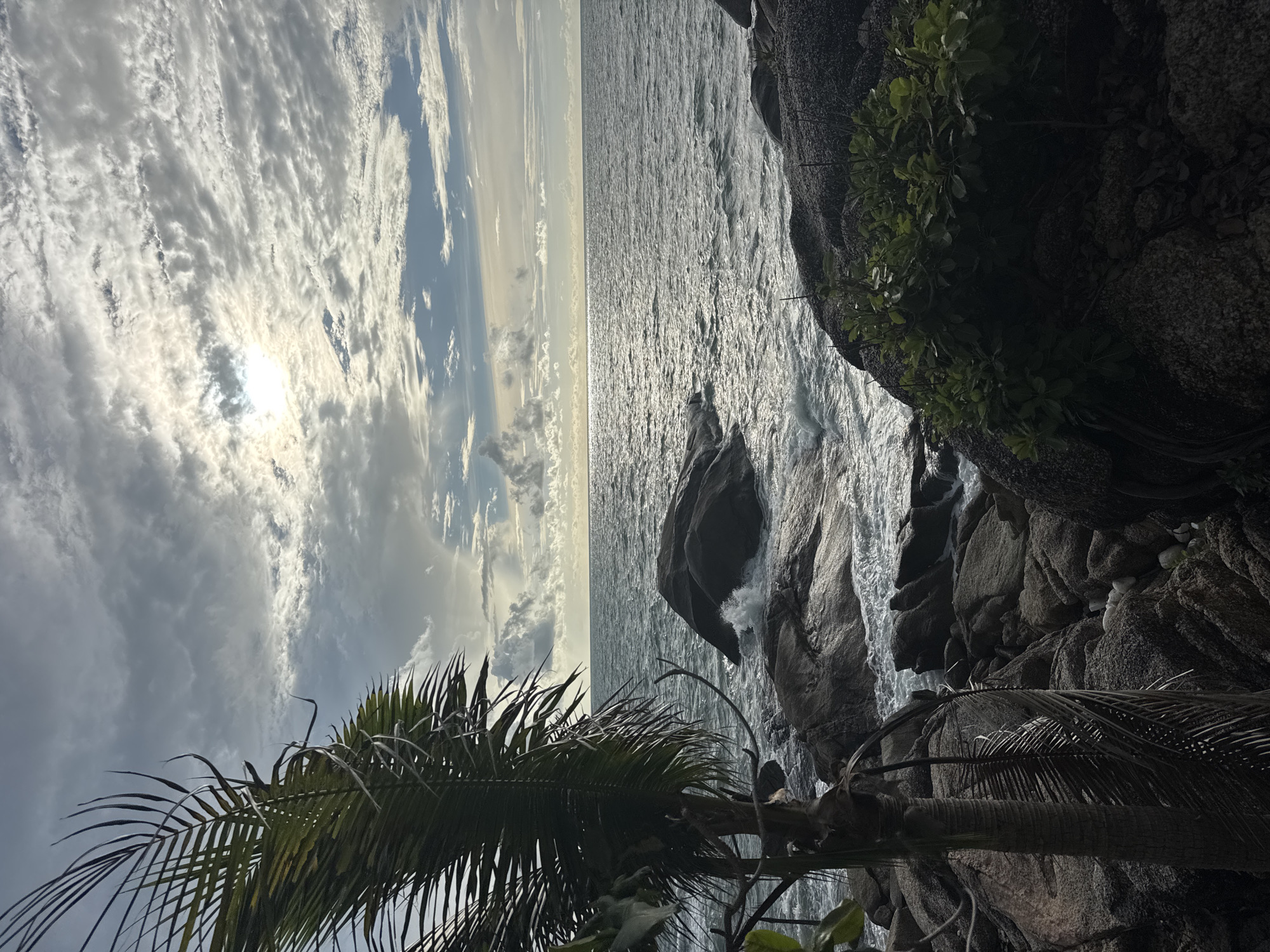 The image shows a coastal view in Baan Thong Nang, Thailand, featuring a rocky shoreline on the right with vegetation. The ocean dominates the center, reflecting sunlight. The sky, filled with striated clouds, is on the left, with the sun partially obscured. A palm tree is visible in the lower left.