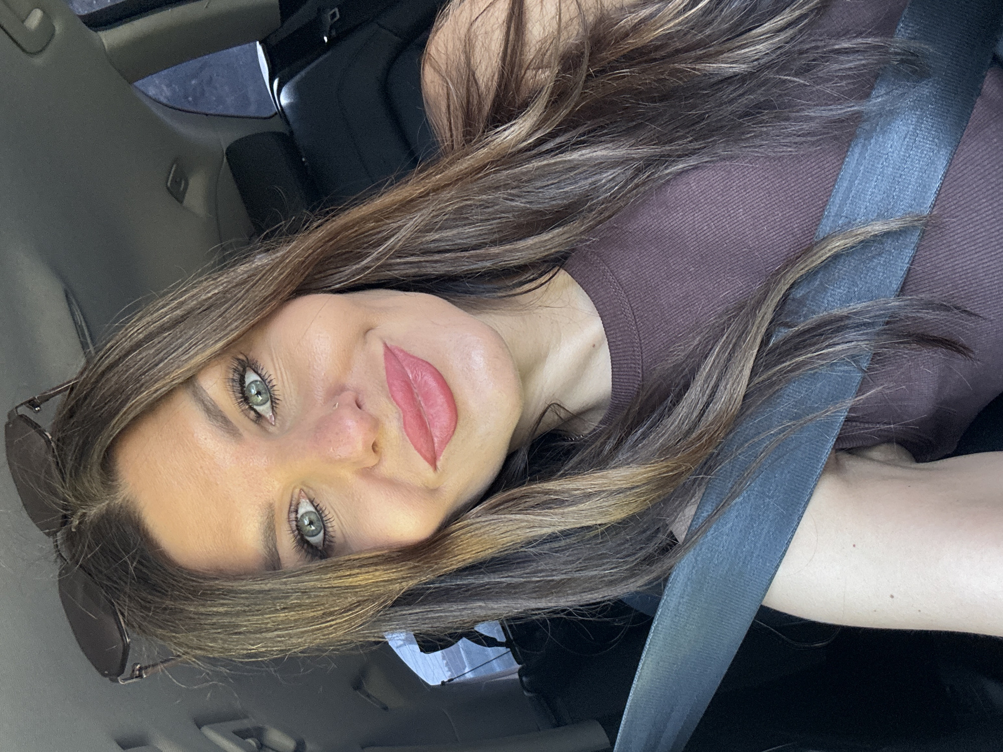 In Noviy, Russia, a woman with long, brown hair and green eyes is taking a selfie in a car. She is wearing a brown top and a seatbelt is across her chest. Sunglasses are perched on top of her head. The interior of the car, including the headrest and the ceiling, is visible.