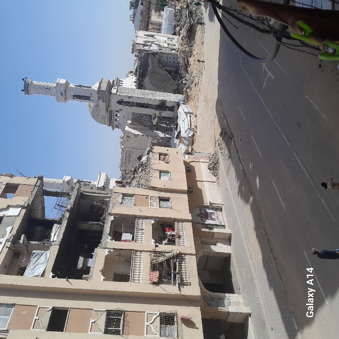 The image depicts a street scene in Khan Yunis, Palestinian Territory, characterized by significant structural damage under a clear blue sky.
In the foreground and midground on the left, a multi-story, light-colored building stands partially destroyed. Multiple floors exhibit exposed interiors, missing walls, and visible rebar. Balconies on the lower levels contain hanging laundry. The ground floor features an arched entrance and a storefront with partial signage. A large white tarp covers a section of a damaged upper floor.
To the right, a heavily damaged white mosque with a tall minaret dominates the background. The mosque's dome is visible, but large sections of its surrounding structure have collapsed into a field of rubble. The minaret itself remains largely upright but shows signs of damage. Piles of debris, including collapsed building materials and scattered white tarpaulins, extend from the mosque's base towards the street.
The foreground features an asphalt road with faded white lane markings. A paved sidewalk borders the damaged building on the left, transitioning to dirt and rubble nearer the mosque on the right. Two individuals are visible on the street and sidewalk in the lower left: one person in dark clothing walks on the sidewalk, followed by another in lighter clothing closer to the road. Further in the background, near the mosque, a few indistinct figures are observable amidst the rubble. Cables or wires are present in the extreme right foreground, suggesting the image was captured from an elevated position.