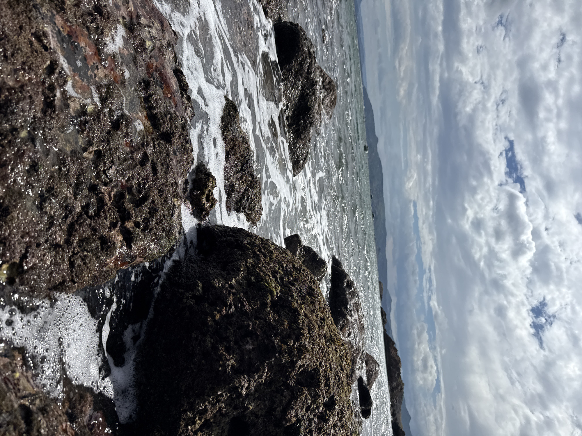 The image depicts a rocky shoreline with the sea lapping at the rocks. In the foreground, dark, rough-textured rocks are partially submerged in foamy, white water. The water reflects the light from the sky. In the middle ground, more rocks are visible, with the sea stretching out to the horizon. The water appears choppy, with sunlight glinting off the surface. In the background, a hazy coastline with hills can be seen, and a few distant boats are dotted on the water. The sky is filled with dramatic, grey clouds, with patches of blue sky peeking through. The scene is likely located on the coast of Song Cau, Vietnam, a region known for its scenic beaches and rocky outcrops.