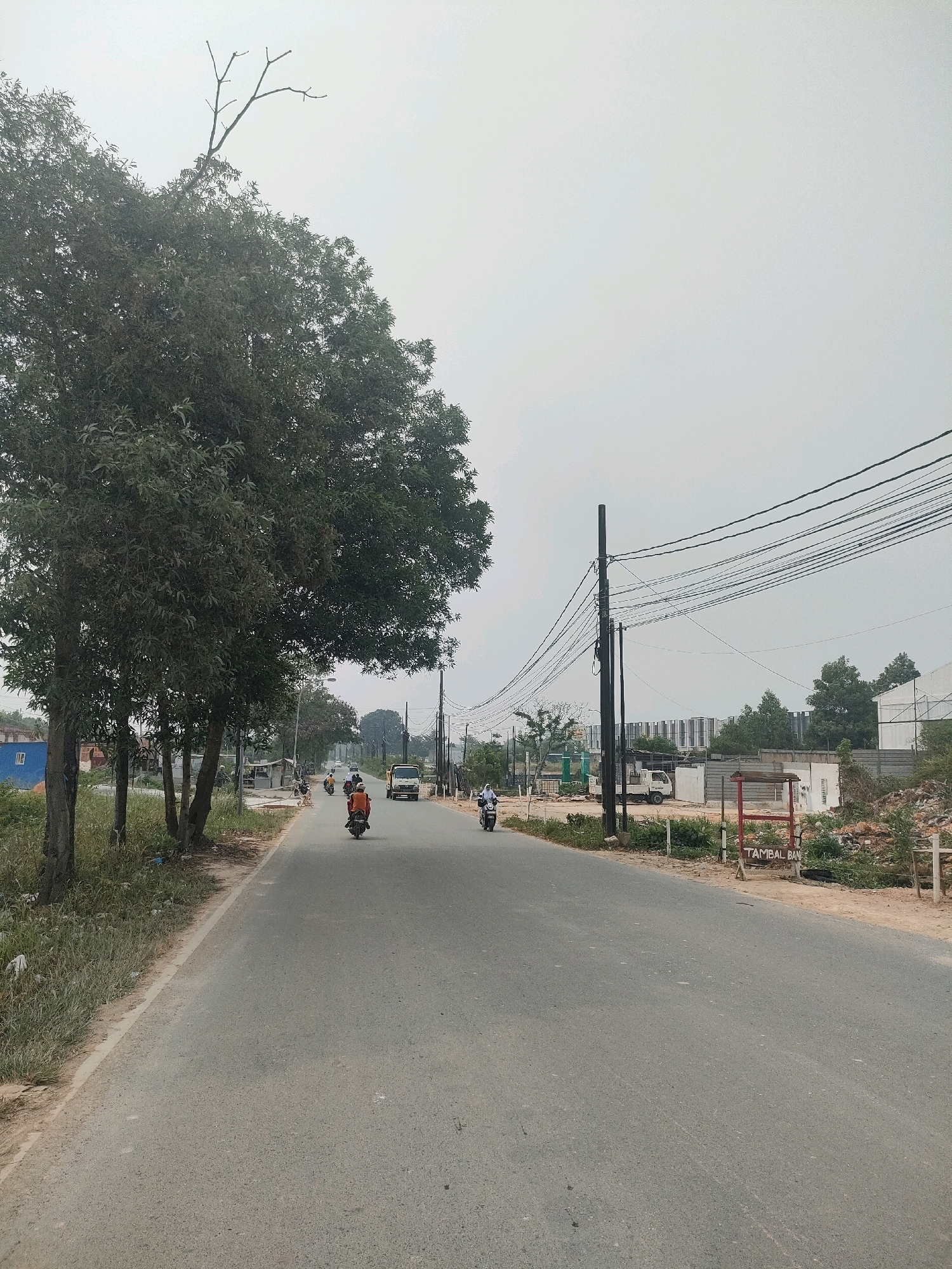 The image depicts a road scene in Batam, Indonesia, under an overcast sky. A paved road stretches into the distance, bordered by a grassy verge and trees on the left. On the right, utility poles with numerous wires are prominent. Several motorcycles are seen traveling on the road, with some riders visible. Further down the road, a yellow truck is present, along with buildings and construction activity. A red sign with white lettering, reading "TAMBAL BAN," indicating a tire repair service, is visible on the right side of the road. The environment appears to be a mix of natural elements and urban development, with ongoing construction suggesting growth in the area.