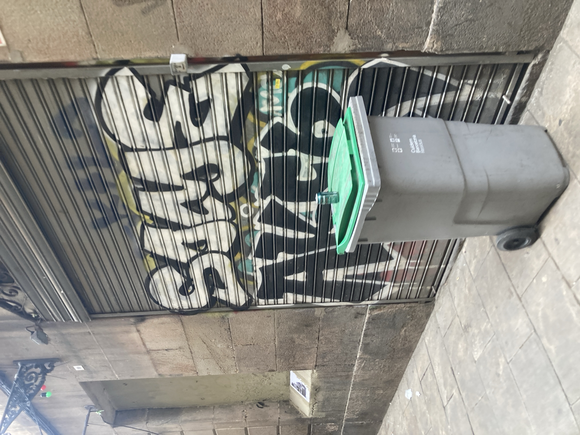 The image shows a grey plastic trash bin with a green lid in front of a closed metal security shutter that is covered in graffiti. The graffiti is large and predominantly in black and white with some yellow and teal accents. The shutter is on a building with a stone facade. To the left of the shutter is a textured stone wall with a recessed niche containing a small framed picture. Above the niche, a metal fixture with two colored lights (green and red) is attached to the wall. The ground in front of the bin and shutter is paved with light-colored square tiles. A single aluminum can sits on the lid of the trash bin. The trash bin has text on its side that appears to read "Ajuntament Barcelona" and "Residus". The scene is likely a street in Barcelona, Spain, given the text on the trash bin.