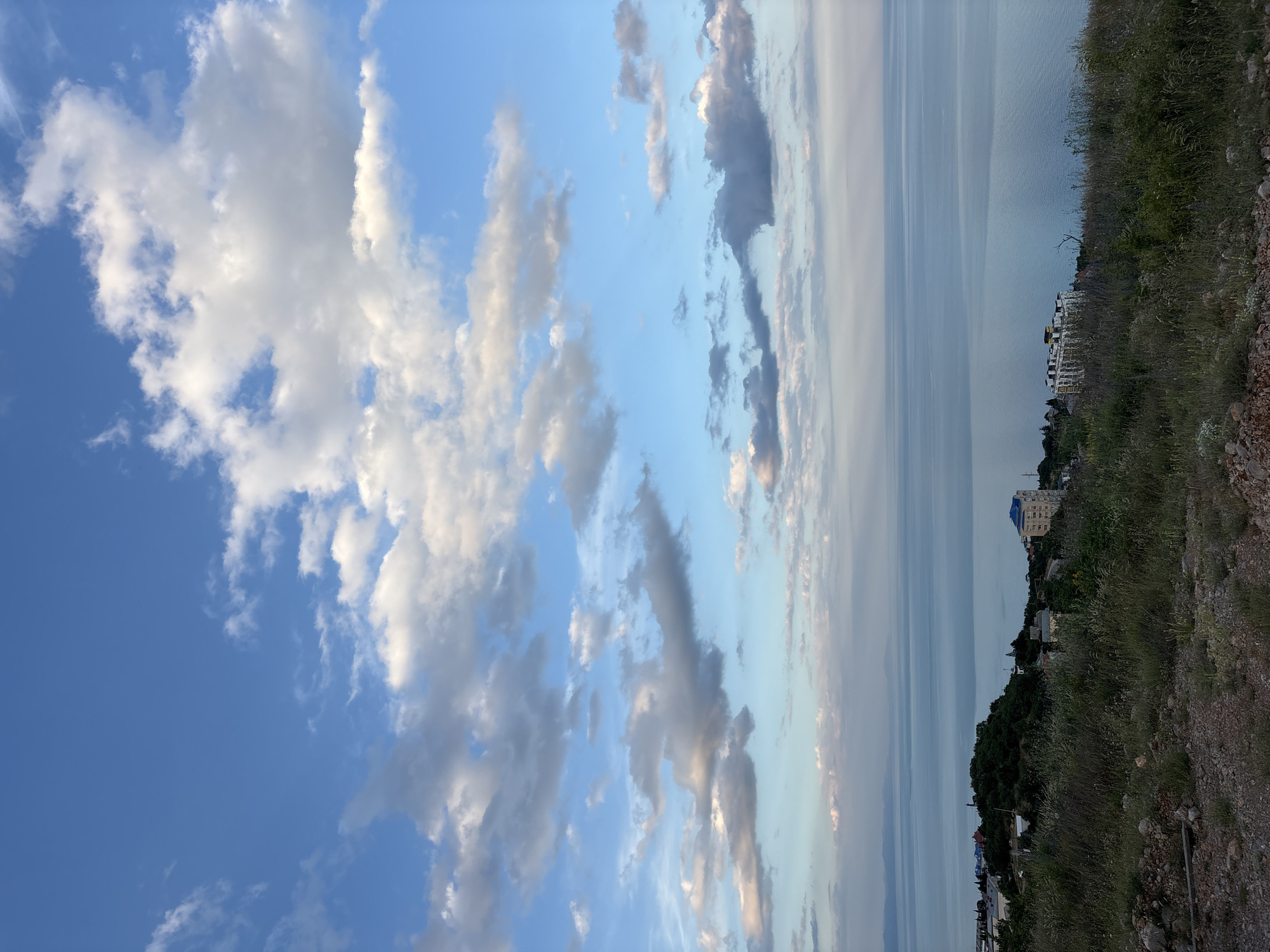 This is a landscape image of a coastal scene with a focus on the sky and the distant sea. The image is oriented vertically. The sky is a prominent feature, filled with a mix of white and gray clouds against a bright blue background, suggesting fair weather. Towards the right side of the image, the sky transitions to a hazy, lighter tone as it meets the horizon.
The sea stretches out in the background, appearing calm and reflecting the muted colors of the sky. Along the coastline, there is a green, tree-covered hillside dotted with buildings. One notable building is a multi-story structure with a light-colored facade and a dark blue roof. Further down the coast, other buildings and structures are visible, nestled amongst the trees and on the slopes. The foreground on the right side of the image is a rocky, overgrown area with sparse vegetation and dry grass, indicating a natural, untamed terrain.
The lighting suggests it is either early morning or late afternoon, given the soft quality of light and the presence of clouds. There is no discernible activity or event taking place; it appears to be a still, observational shot of the natural environment. No text is visible in the image. The scene evokes a sense of tranquility and the vastness of nature.