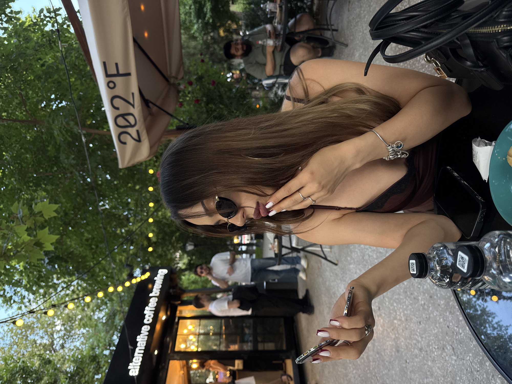 In an outdoor seating area of a cafe, a woman with dark brown hair, wearing sunglasses and a maroon top, checks her reflection in a compact mirror. She has burgundy lipstick on and her nails are painted with a white French manicure. She wears a bracelet with charms on her left wrist and several rings on her fingers. A black handbag is visible to her right. On the table in front of her are two plastic water bottles and a smartphone. In the background, a man in a green shirt is seated at a table. Behind him, two men stand near a building with a sign that reads "alternative coffee brewing." Overhead, string lights are visible amidst lush green trees, suggesting a pleasant, possibly summer, day. The umbrella above the woman is imprinted with "202°F". The overall scene suggests a relaxed outdoor dining or coffee experience in Yerevan, Armenia, likely during daylight hours.