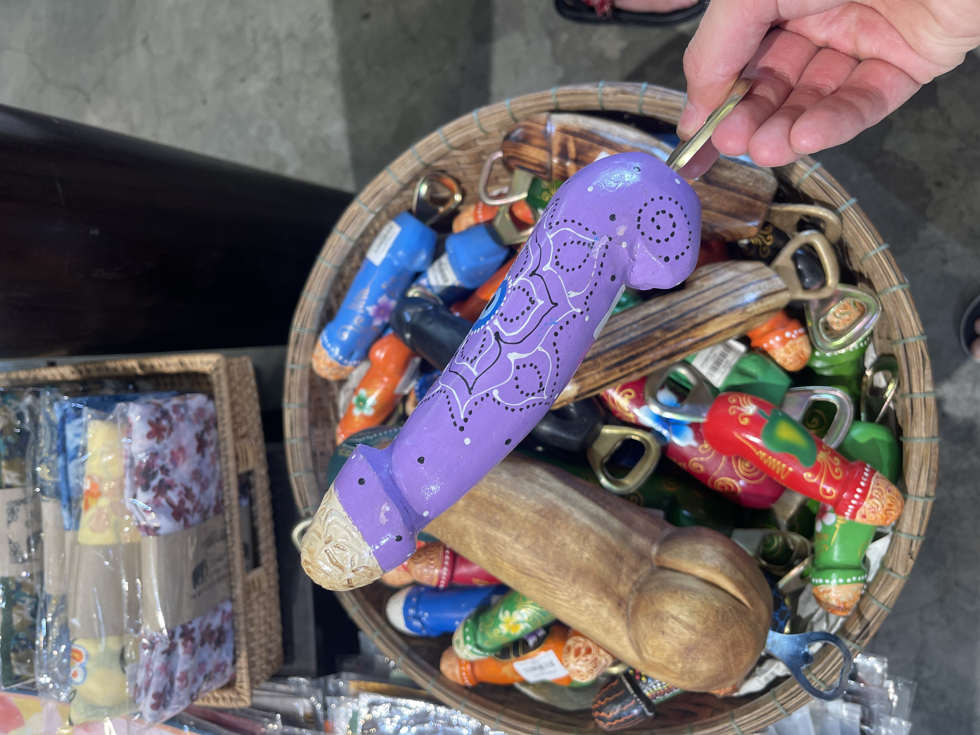 The image shows a hand holding a purple, phallus-shaped bottle opener over a basket filled with similar bottle openers of various colors and materials, including wood, in what appears to be a market stall. The basket sits on a surface next to a woven basket containing packaged goods with floral patterns. The location is Nha Trang, Vietnam.