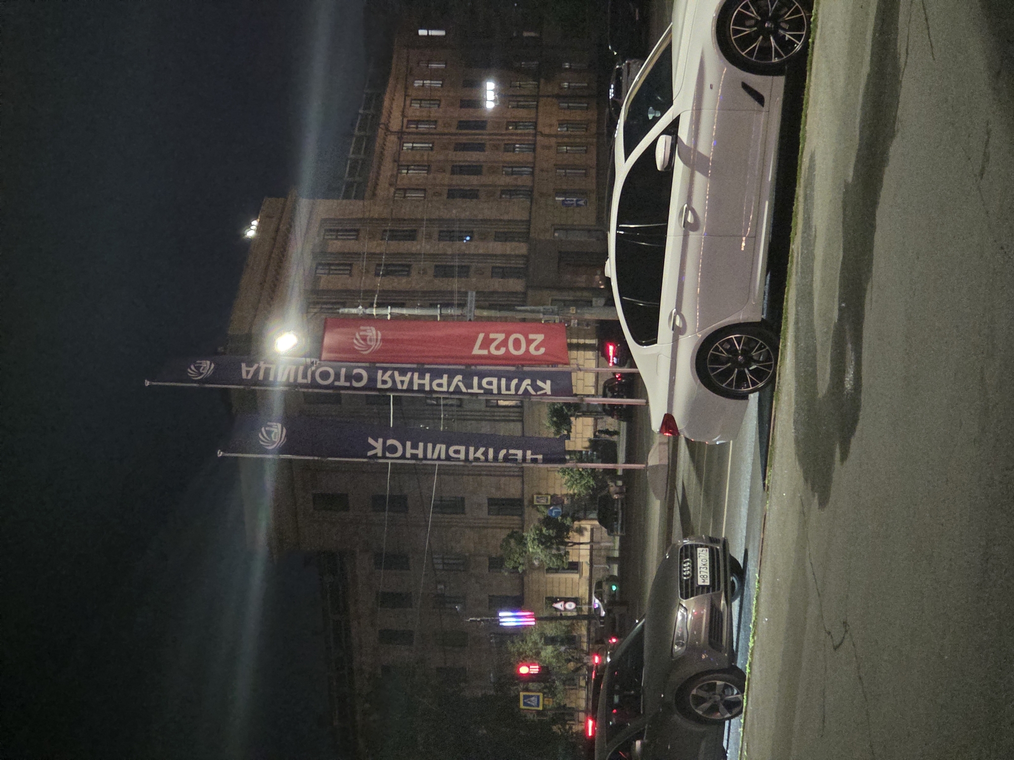 At night, on a city street in Chelyabinsk, Russia, two cars, a dark sedan and a white sedan, are parked facing left. The dark sedan, an Audi, has its license plate visible, reading "M873KO 775". In the background, a multi-story building with lit windows stands under a dark sky. Several tall flags are erected in front of the building. Two of the flags are dark blue with white text, and one is red. The blue flags appear to have text reading "ЧЕЛЯБИНСК" (Chelyabinsk) and "КУЛЬТУРНАЯ СТОЛИЦА" (Cultural Capital). The red flag has "2027" printed vertically. Traffic lights and streetlights are also visible. The street is wet, suggesting recent rain or a damp surface.