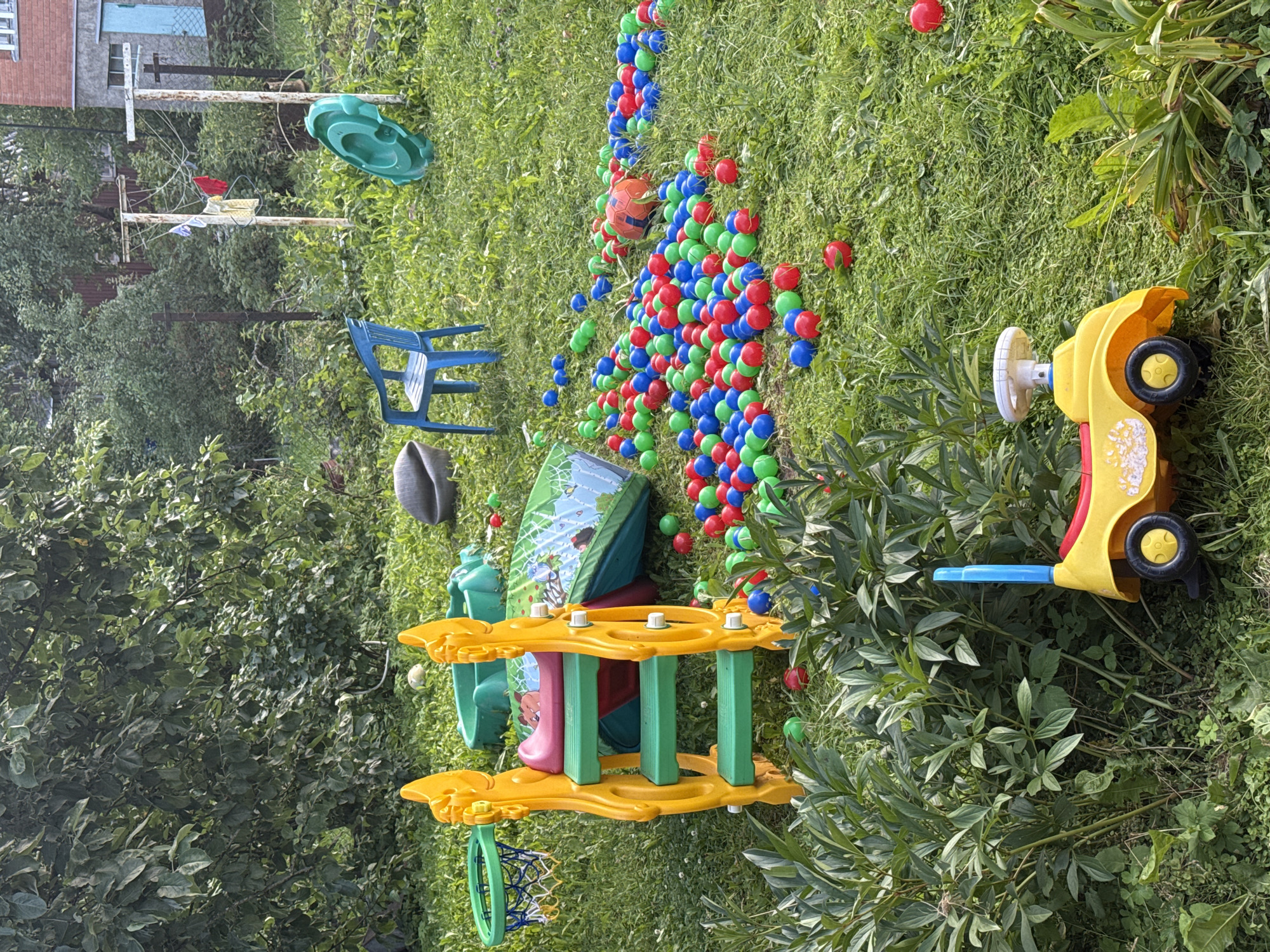 The image depicts a cluttered backyard in Дзержинец-83 Снт, Russia, filled with children's toys and overgrown vegetation. A yellow plastic slide with green steps is centrally located, accompanied by a blue plastic chair, a green plastic roundabout, and a basketball hoop. A large pile of colorful plastic balls, predominantly red, green, and blue, is scattered across the grass, along with an orange ball. A yellow toy ride-on car with a blue handle is positioned in the foreground. The environment appears unkempt, with tall grasses and leafy plants encroaching on the play area. In the background, a clothesline with laundry is visible, along with a portion of a brick building. The overall scene suggests a disused play area in a rural or suburban setting.