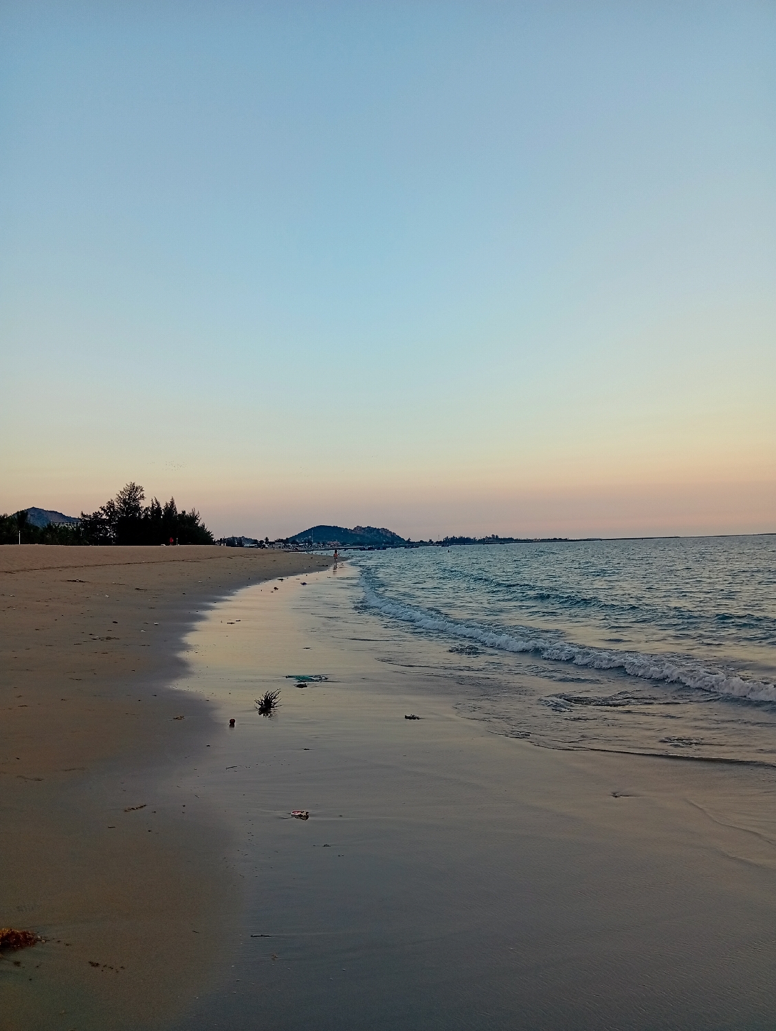 The image depicts a serene coastal landscape during what appears to be either sunrise or sunset, characterized by the soft, gradient colors of the sky. The upper sky transitions from a clear, light blue to pale orange and pink tones closer to the horizon.
A wide sandy beach dominates the foreground and extends into the distance on the left side of the frame. The sea occupies the right side, with gentle waves breaking onto the shore, leaving foamy white lines and wetting the sand, which reflects the sky's soft light. Scattered pieces of natural debris, such as dark clumps of seaweed, are visible on the wet sand.
Along the distant left coastline, trees and vegetation are visible, leading to a series of low hills or mountains. Similar mountainous landforms can be seen further in the distance across the water on the right horizon. A few small, indistinct human figures are present in the far distance; one appears to be walking on the wet sand near the waterline, and another is possibly in the shallow water.
The overall scene conveys a sense of calm and tranquility. No visible text is present in the image. The setting is a beach in Khánh Hải, Vietnam.