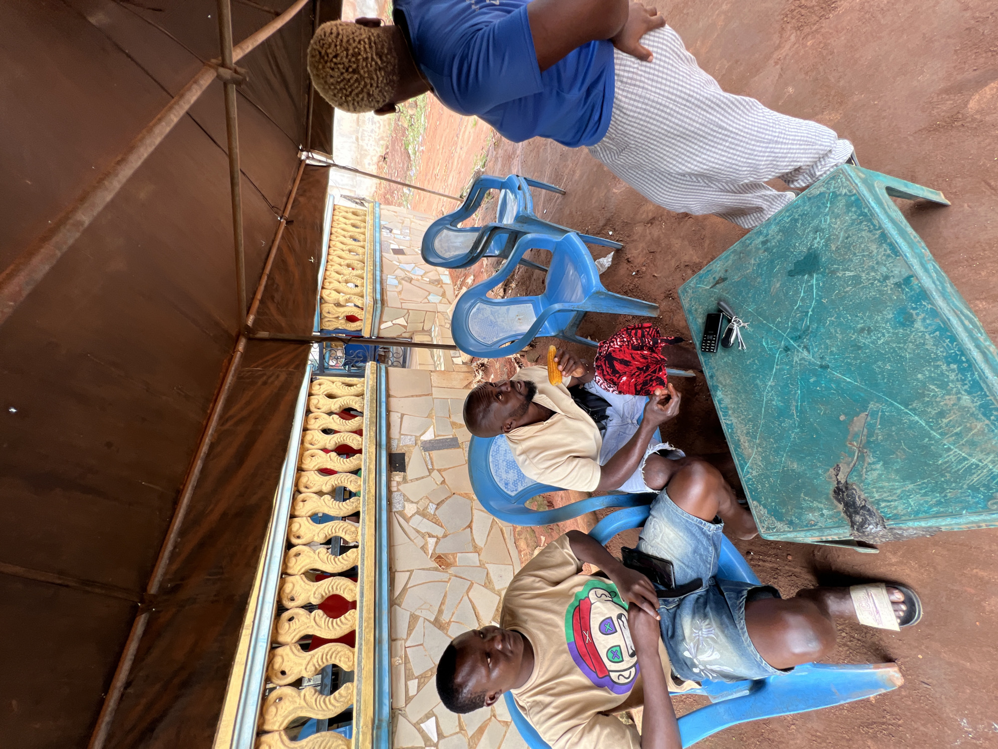 In Nkpor, Nigeria, under a brown tarpaulin roof supported by metal poles, three individuals are gathered around a teal-colored table. The ground is covered in reddish-brown dirt. Two men are seated on blue plastic chairs. The man on the left, wearing a tan t-shirt with a graphic print and denim shorts, looks towards the right. The man in the middle, in a light tan short-sleeved shirt and denim shorts, holds a piece of corn in his right hand and a red and black patterned cloth in his left. A woman with blonde, curly hair, wearing a blue t-shirt and striped pants, stands facing away from the camera, her hands on her hips. Behind the seated individuals is a wall with a decorative yellow and blue balustrade and sections of mosaic tiling. A dark brown, potentially metal, bar runs along the top of the balustrade. A mobile phone and keys are visible on the table.