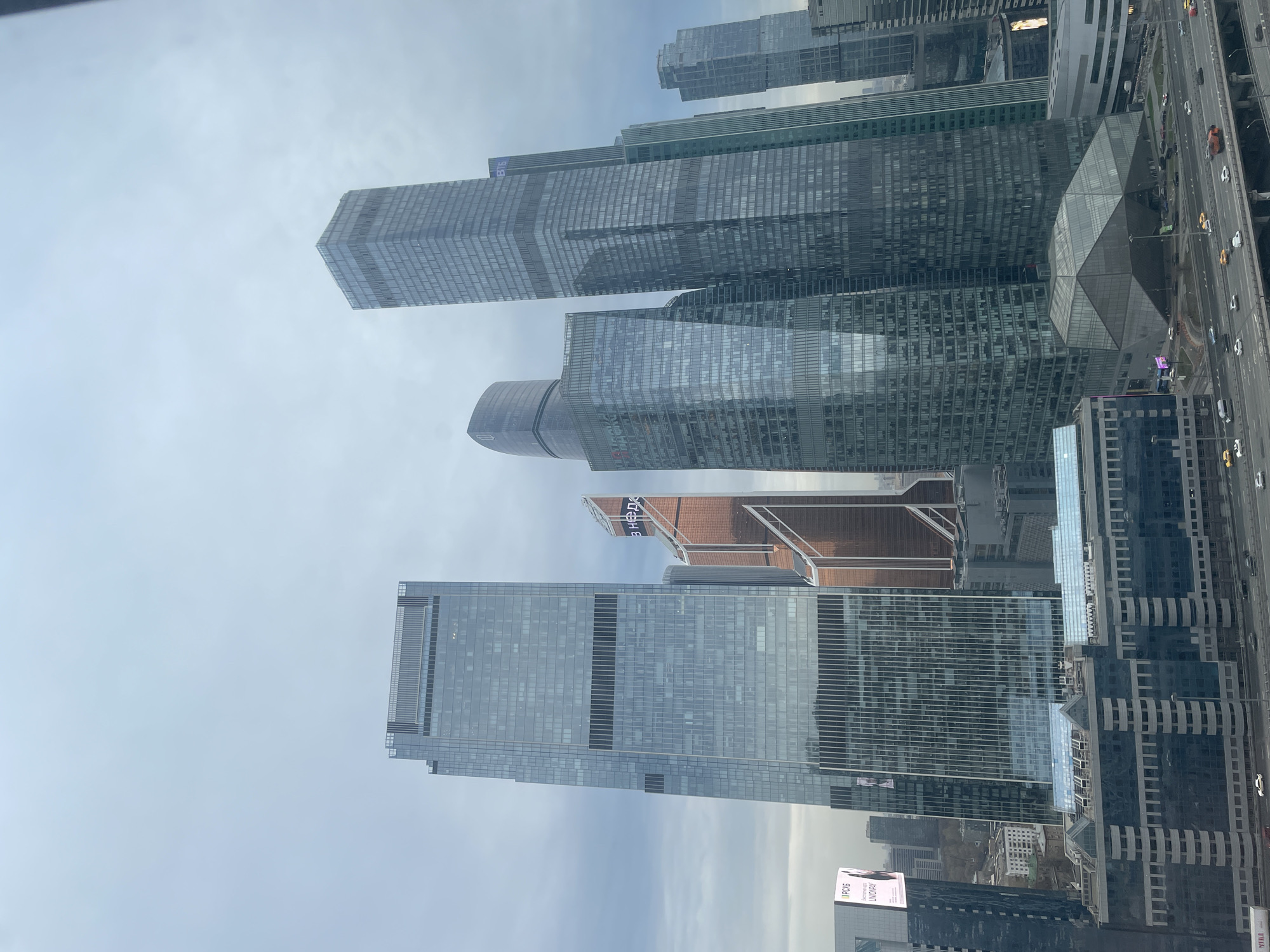 This image shows an upward-looking view of a cluster of modern high-rise buildings in a city, presumably within Khimki, Russia. The buildings are characterized by glass facades and geometric shapes. The road and vehicles below are only visible on the right. Overcast sky in the background.