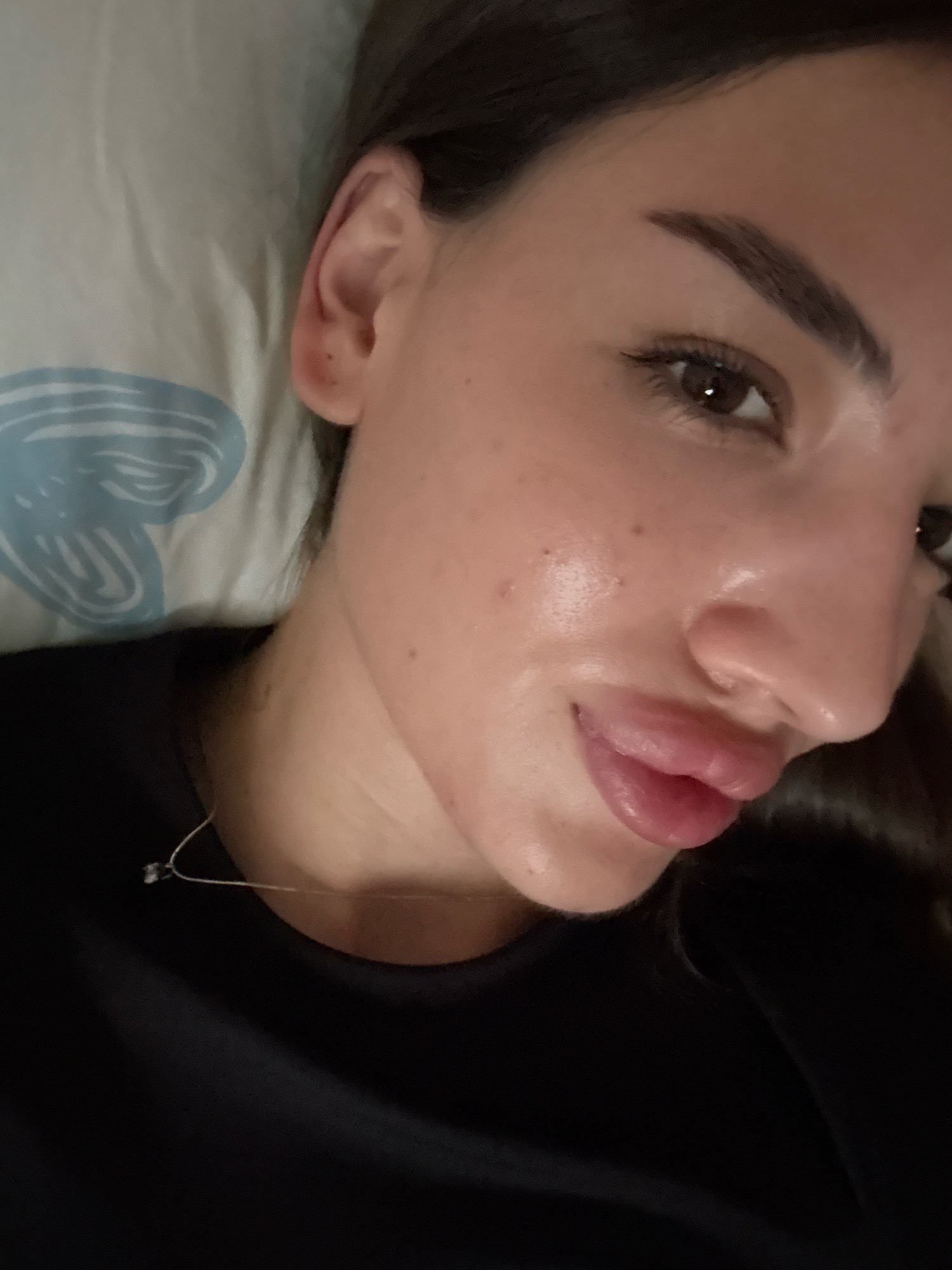 A close-up, high-angle, indoor shot features a young woman lying on her side, facing the camera. She has dark hair pulled back, and her face is illuminated, highlighting her smooth skin, full lips, and well-defined eyebrows. Her right eye is visible, with a subtle winged eyeliner. She is wearing a black t-shirt, and a thin, silver chain with a small pendant rests on her collarbone. The background consists of a pillow with a light blue abstract design. The scene appears to be a bedroom or a comfortable indoor setting, possibly in Basarabeasca, Moldova, given the context.