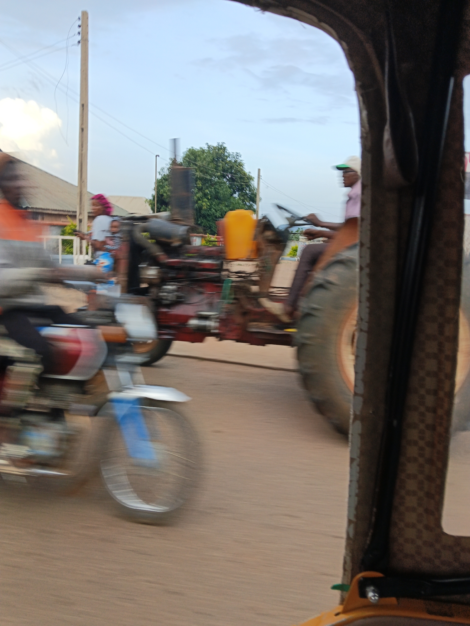 This image depicts a street scene in Bukuru, Nigeria, captured during daytime under a cloudy sky. In the foreground, a motorcycle is visible, with its front wheel blurred due to motion. A person is seated on the motorcycle, wearing dark clothing. To the right, a tractor is also in motion, with a person driving it, wearing a pink shirt and a green cap. The tractor has large rear wheels and a yellow tank on its side. A power pole with wires is present in the background, along with some trees and buildings. The overall impression is that of a busy, active street with vehicles and pedestrians. The motion blur suggests that the photograph was taken while the camera was moving.