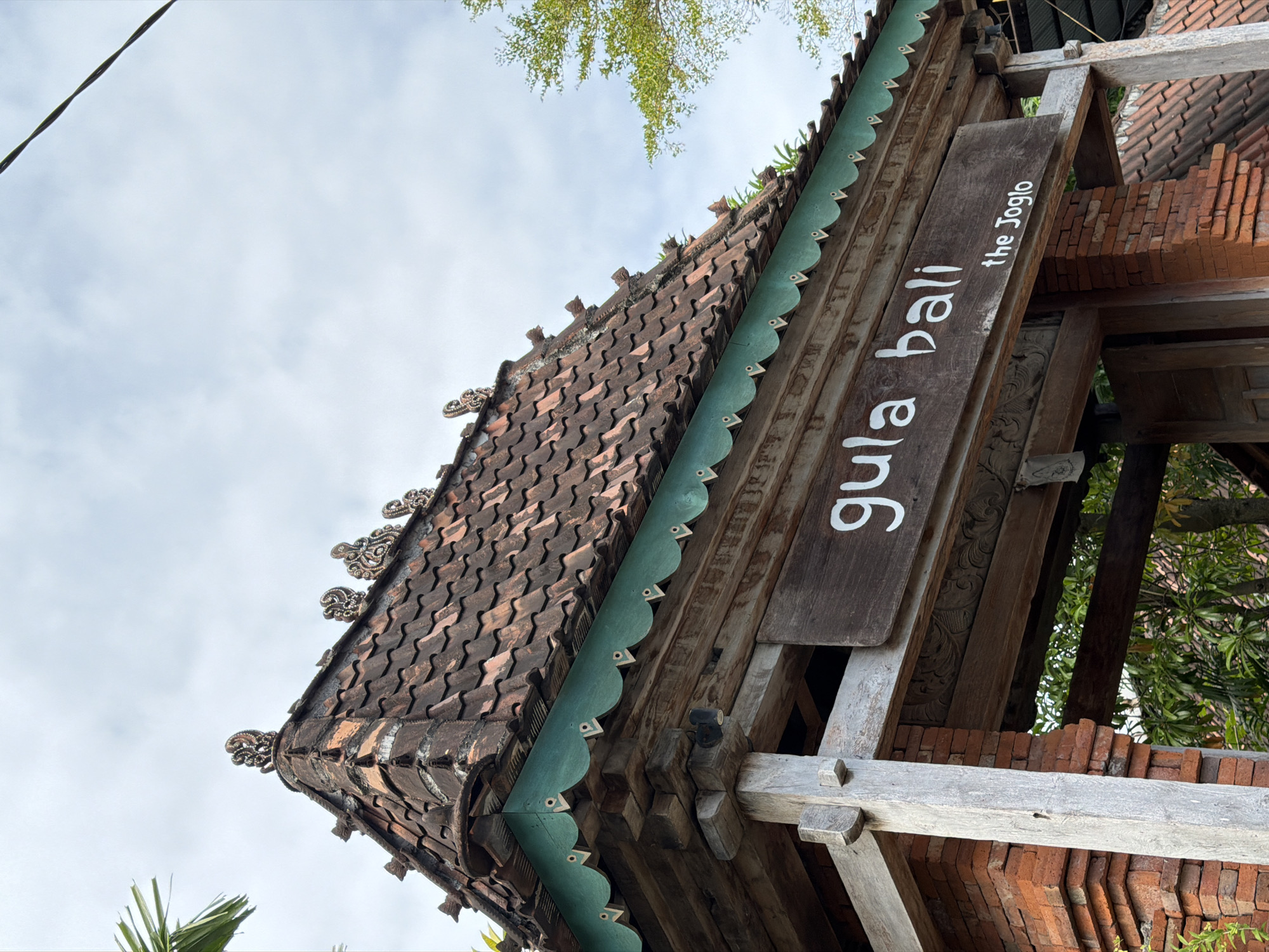 The image captures a detailed view of an outdoor architectural feature, likely an entrance or a signage area, in Kuta, Indonesia. The focal point is a weathered wooden sign with white lettering that reads "gula bali" on the top line and "the Joglo" on the bottom line. This sign is mounted on a wooden beam that forms part of a larger structure.
The structure itself is characterized by traditional Balinese architectural elements. A steeply pitched roof covered with terracotta tiles is visible, with decorative finials adorning the ridge. Below the roof, a band of intricately carved wooden trim painted a vibrant green runs along the eaves, featuring a scalloped edge and triangular decorative elements. The supporting pillars are made of roughly hewn, weathered wood, and the walls are constructed from reddish-orange bricks, showcasing a rustic and aged aesthetic.
The surrounding environment suggests a tropical setting. Green foliage is visible through the openings of the structure, and the sky above is overcast with clouds. A dark electrical wire diagonally crosses the upper portion of the frame. No people or animals are present in the image. The overall impression is one of historical charm and cultural authenticity.