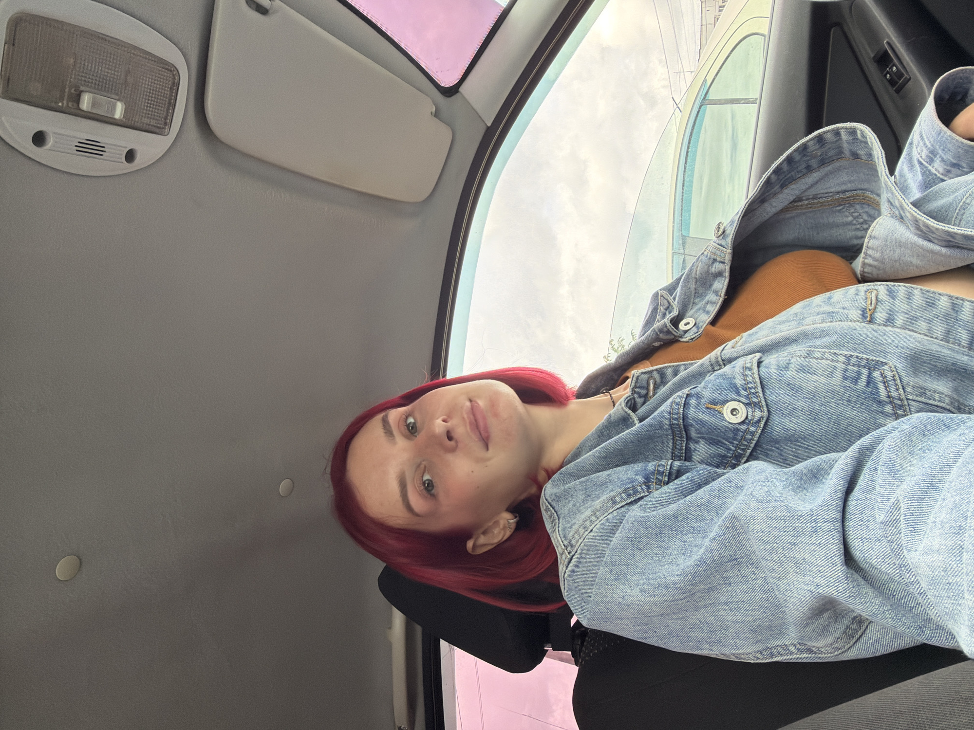A young woman with distinctive bright red hair is captured in a selfie or portrait, seated inside a car in Chaikovsky, Russia. The image is oriented with the woman's head to the left, looking directly at the camera with a neutral to slightly pleasant expression. She is wearing a light blue denim jacket over an orange or earthy-toned top. Her left ear features a small silver piercing.
The setting is the interior of a car, likely from the front passenger seat perspective. Above her, the light gray headliner of the car is visible, along with a dome light fixture and a sun visor. The top of a car seat headrest is visible behind her neck. Through the car window, an overcast or cloudy sky can be seen, suggesting daytime. Some faint outlines of distant structures or trees are also visible beyond the window. The car's door frame and a small control panel on the door are partially visible on the right side of the image. The general atmosphere suggests a casual moment, possibly during travel or while parked. There is no discernible text visible within the image.
