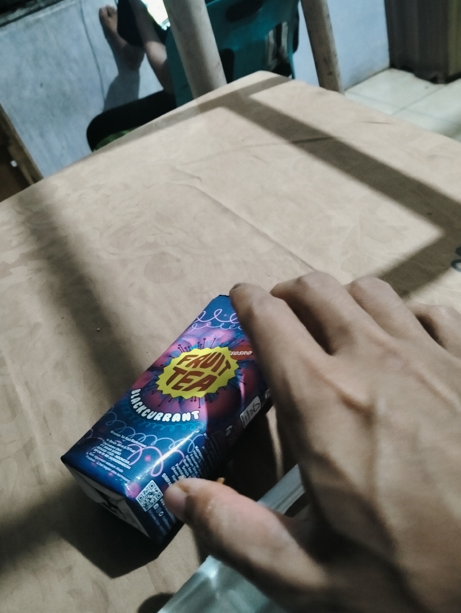 In the City of Medan, Indonesia, a person's hand, appearing to be that of a young adult, is positioned over a beverage can. The can, with a dark blue base and a vibrant, swirling design featuring shades of pink, purple, and yellow, prominently displays the words "FRUIT TEA" in large, bold yellow lettering within a starburst shape. Below this, "BLACKCURRANT" is written in white. The can also features a QR code and other text in small print, likely including product information. The surface beneath the can is a light brown, possibly a table covered with paper or a similar material. In the background, a blurry figure of a person is visible, seated and partially obscured, with shadows cast on a pale blue wall. The overall lighting suggests an indoor setting, possibly at dusk or night, with artificial light sources creating distinct shadows. The focus is on the beverage can and the hand interacting with it, hinting at an act of consumption or engagement with the product.