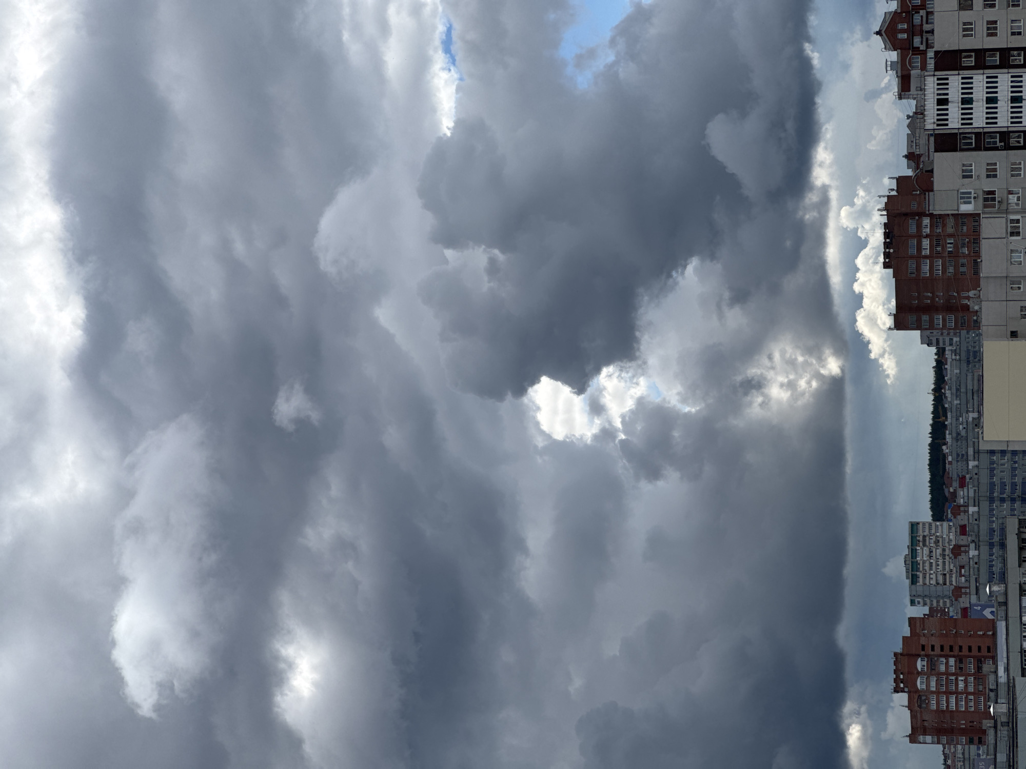 The image presents a dramatic urban scene dominated by a heavily clouded sky. The sky occupies the majority of the frame, filled with dense, dark grey clouds indicative of stormy weather or a powerful weather front. These formidable clouds display varied textures and depths, suggesting significant atmospheric activity.
Towards the upper right of the sky and in several scattered patches, there are visible breaks in the heavy cloud cover, revealing segments of clear blue sky and brighter white clouds. Sunlight appears to penetrate or reflect off certain areas of the clouds, creating bright highlights and enhancing the dynamic, somewhat moody atmosphere.
Along the bottom of the image, a horizon of multi-story buildings defines an urban or suburban landscape. These structures vary in architectural style and color; some are constructed with reddish-brown brick, while others feature lighter grey or white facades with numerous windows. A distant line of trees or a forested area is faintly visible beyond the immediate buildings, marking the edge of the cityscape.
The time of day appears to be daytime, likely mid-day or afternoon, given the discernible light despite the extensive cloud cover. The scene captures a moment of powerful natural weather over a man-made environment. There is no discernible text or human activity in the image.