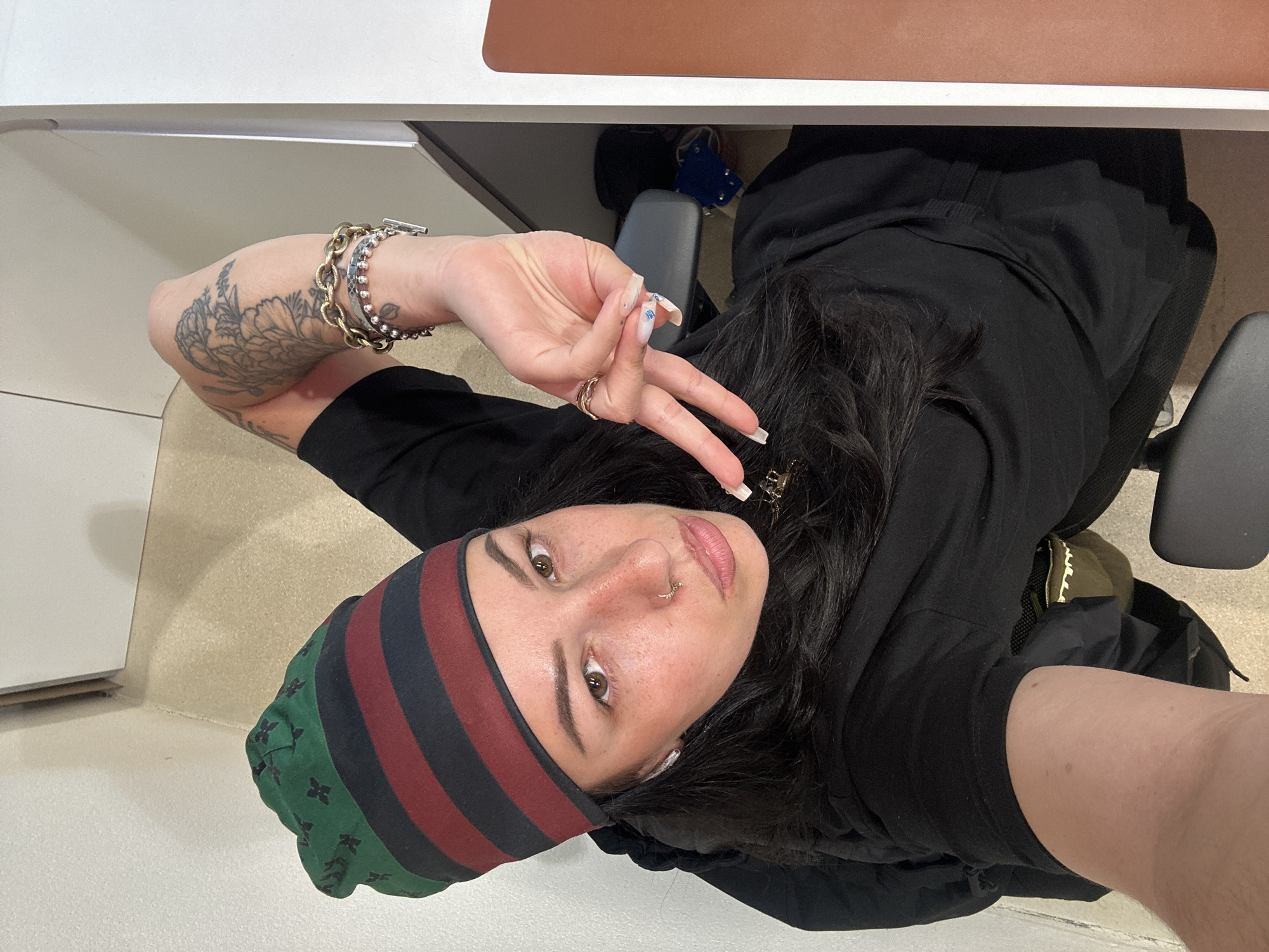 A woman poses for a selfie in an indoor setting. She is wearing a dark-colored top, a striped beanie with green floral accents, and several bracelets on her left wrist. She is making a hand gesture with elongated nails. Her hair is long and dark, and she has a nose piercing. The background includes a desk with a light-colored surface and a chair with gray elements. The overall lighting appears to be bright, and the image is taken from an overhead perspective. The location is not discernable in the city of Paris, France.