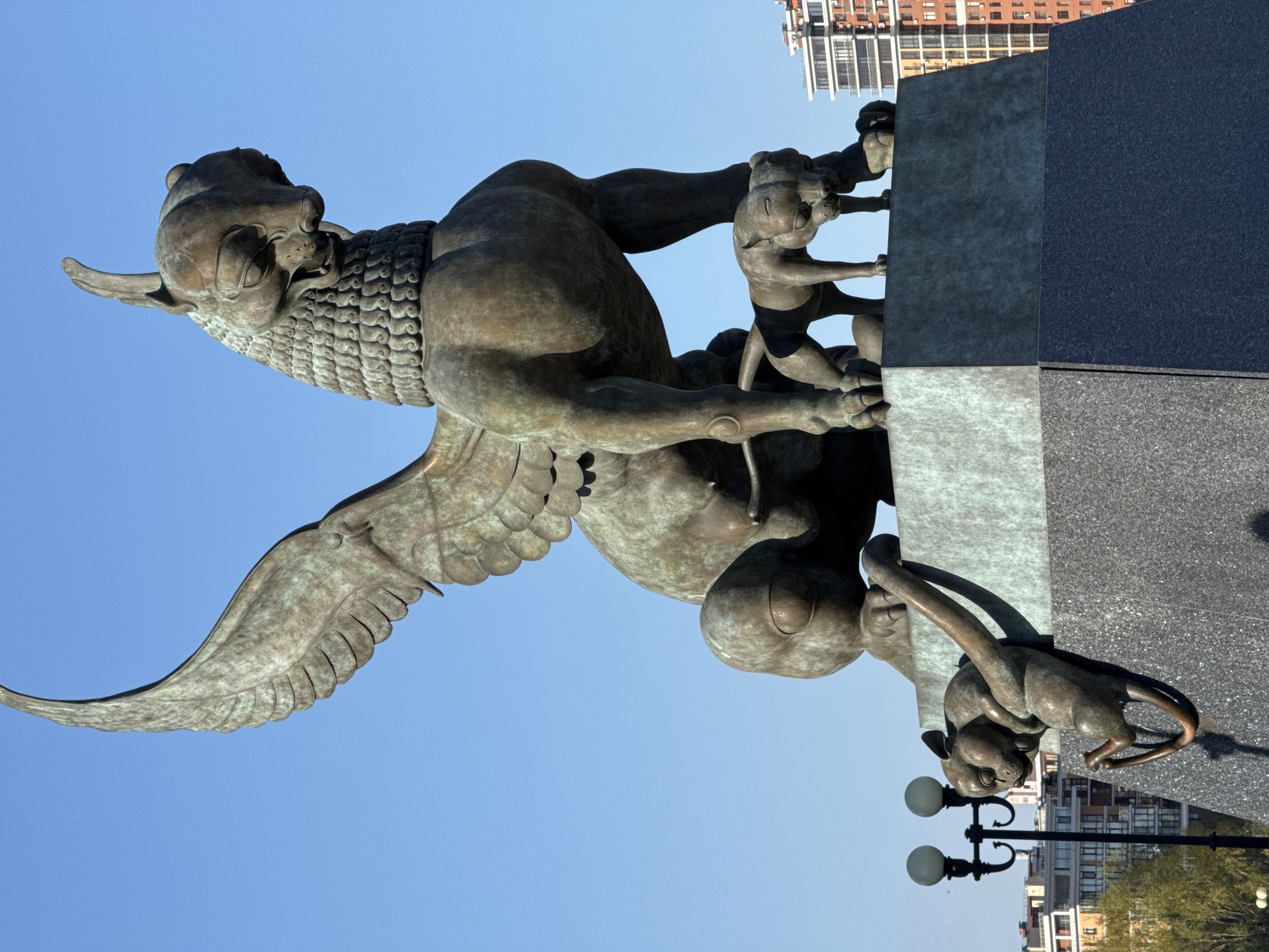 A bronze sculpture in Kazan, Russia, features a winged snow leopard with children clinging to a stone pedestal. The sculpture stands against a clear blue sky, with buildings visible in the background. The pedestal is made of multiple panels of different stone textures. A streetlight is visible near the base of the sculpture.