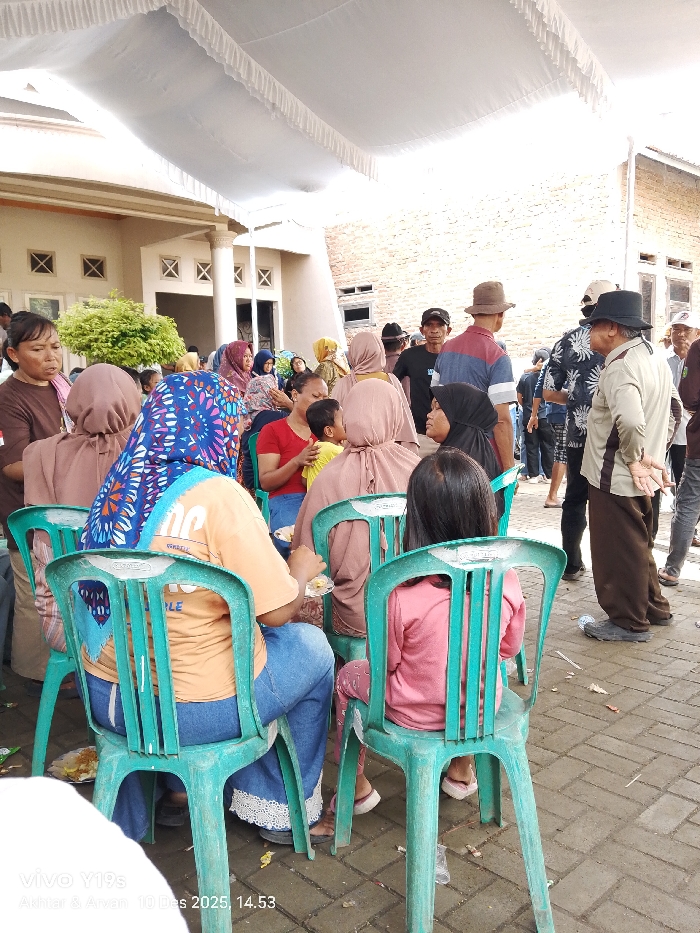 A gathering of people outside, possibly for an event, under a large white awning. Many individuals are seated in rows of green plastic chairs. Several wear head coverings, and some are eating from plates. Others stand around, chatting. In the background, a building with windows and greenery is visible, along with a brick wall. The ground appears to be paved. The scene is captured from a slightly low angle.